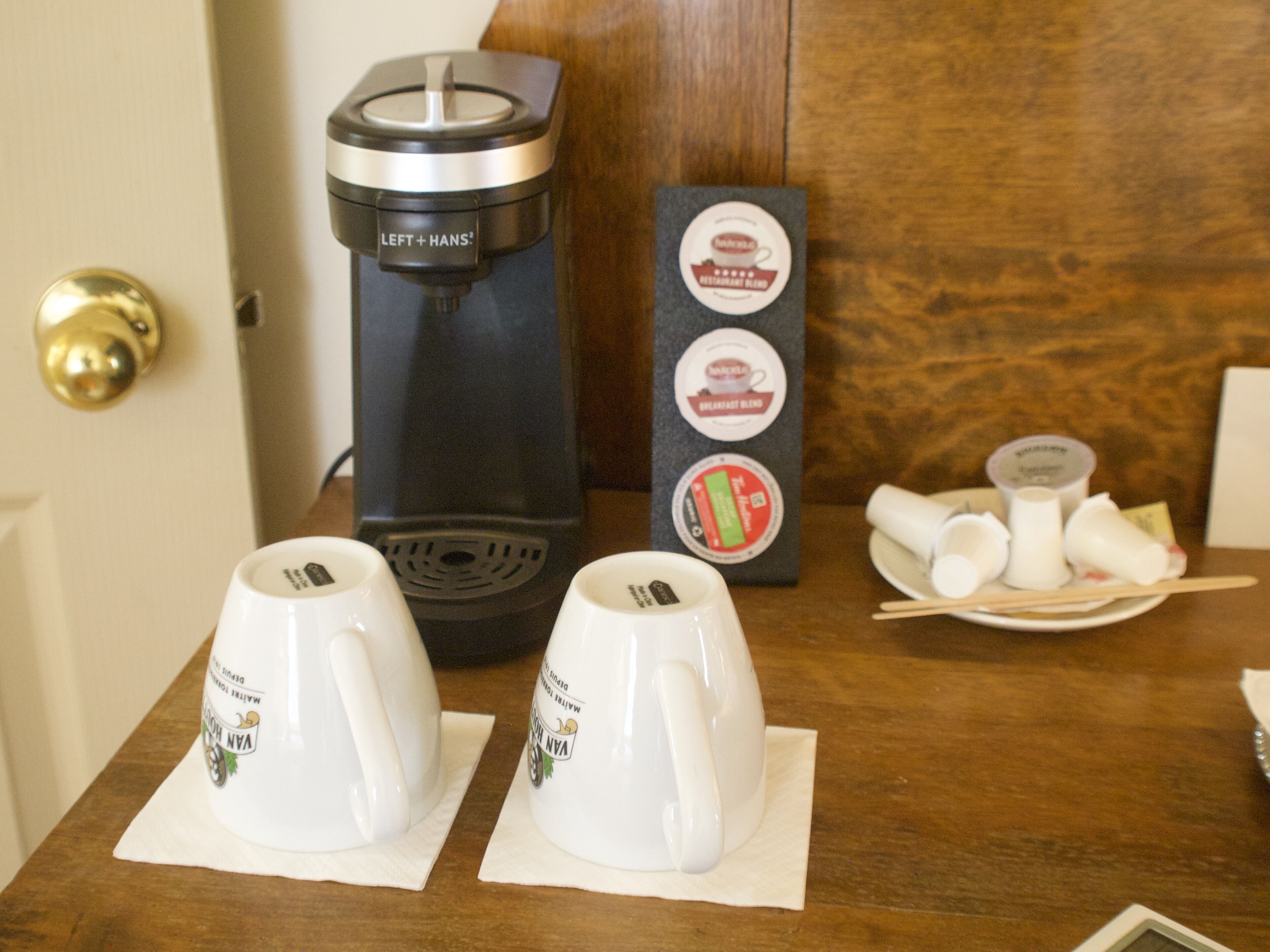 A coffee maker, two white cups, and a napkin on a wooden table.