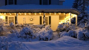 A snow-covered landscape with a house featuring a porch and string lights. - Au Manoir de la rue Merry (Magog)
