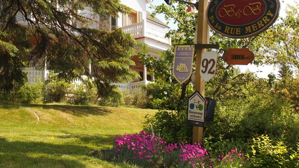 A signpost with a vintage design, a house in the background, and a lush garden with flowers. - Au Manoir de la rue Merry (Magog)