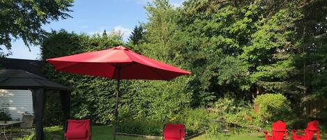 Een terras met rode stoelen en een rode parasol, omringd door weelderige begroeiing en bomen.