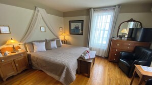 A hotel room with a bed, bedside tables, a desk, a chair, a TV, and a window with curtains. - Au Manoir de la rue Merry (Magog)