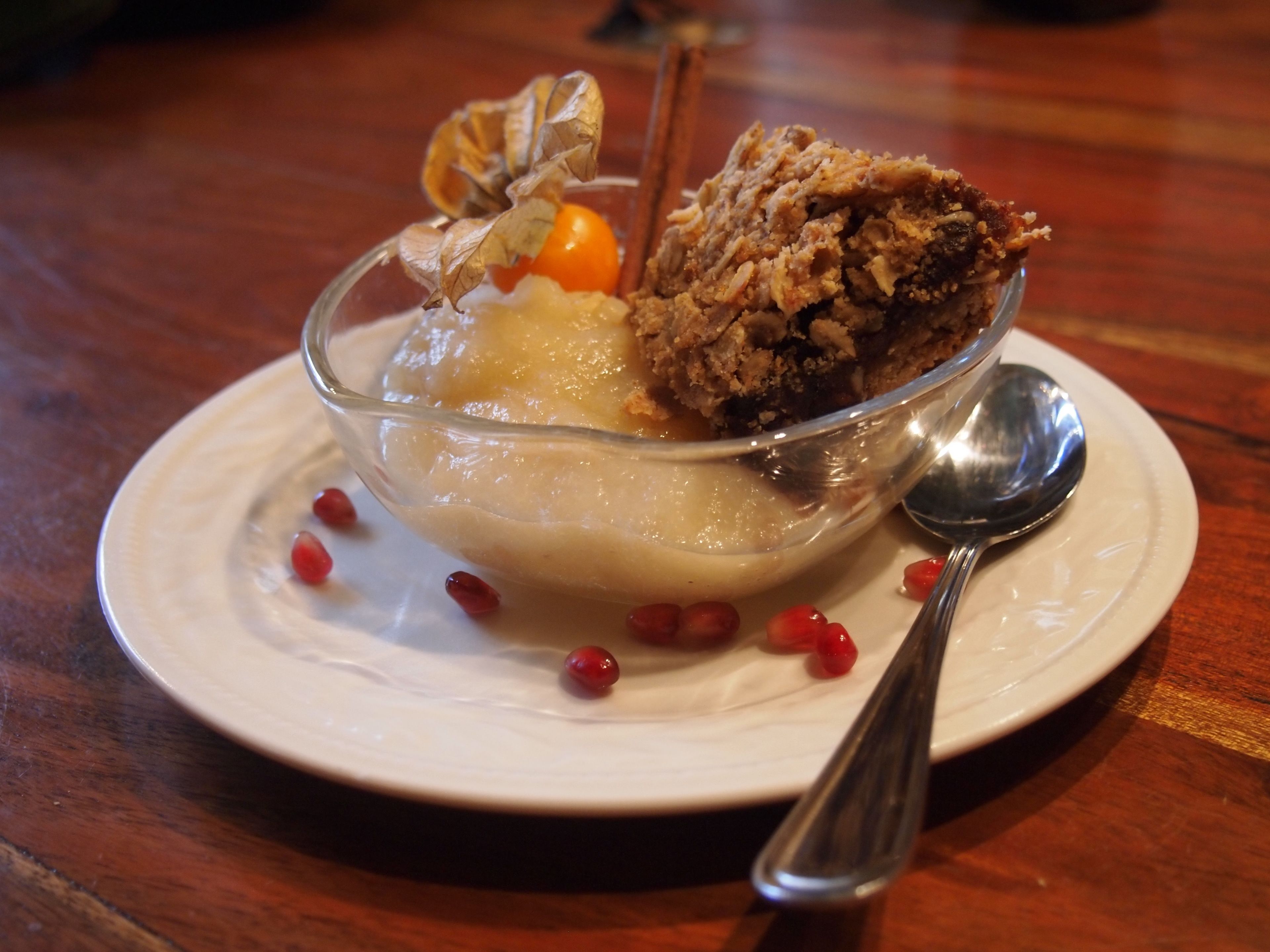 A dessert in a clear glass bowl on a white plate with a spoon, garnished with dried leaves and pomegranate seeds.