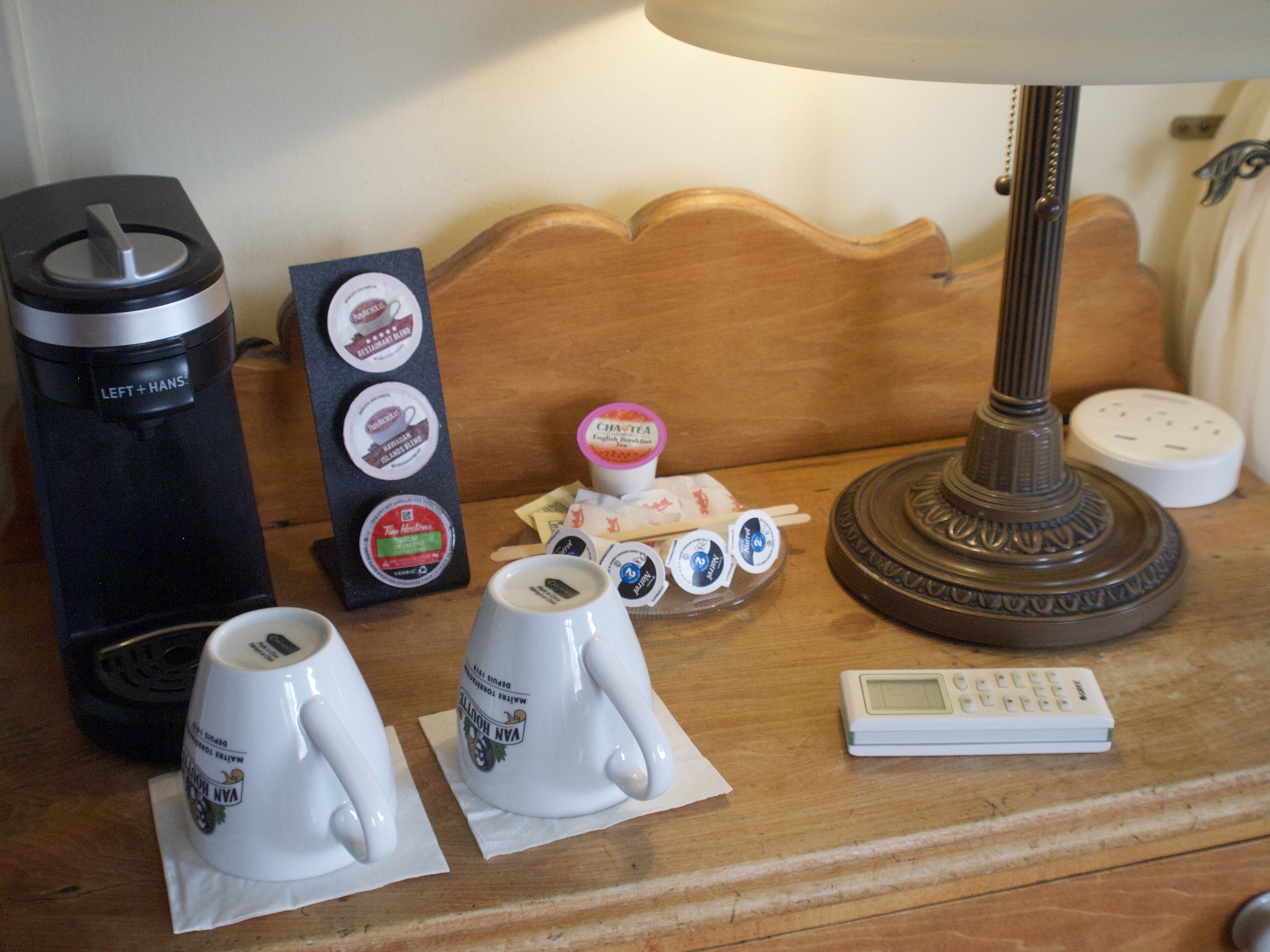 A table with a coffee maker, two coffee cups, a tea cup, a small toy, and a remote control.