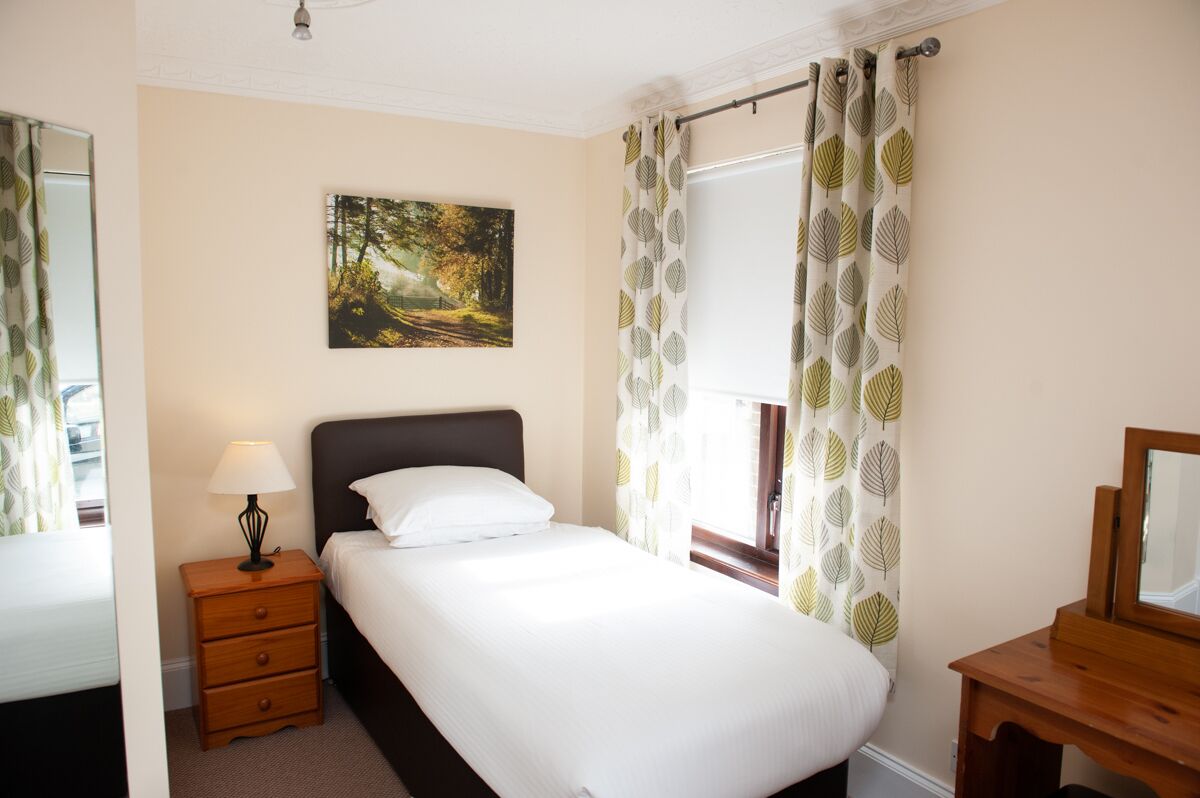 A neatly made bed with a white comforter and pillows, a bedside lamp, a small wooden nightstand, a framed picture on the wall, and a window with leaf-patterned curtains.