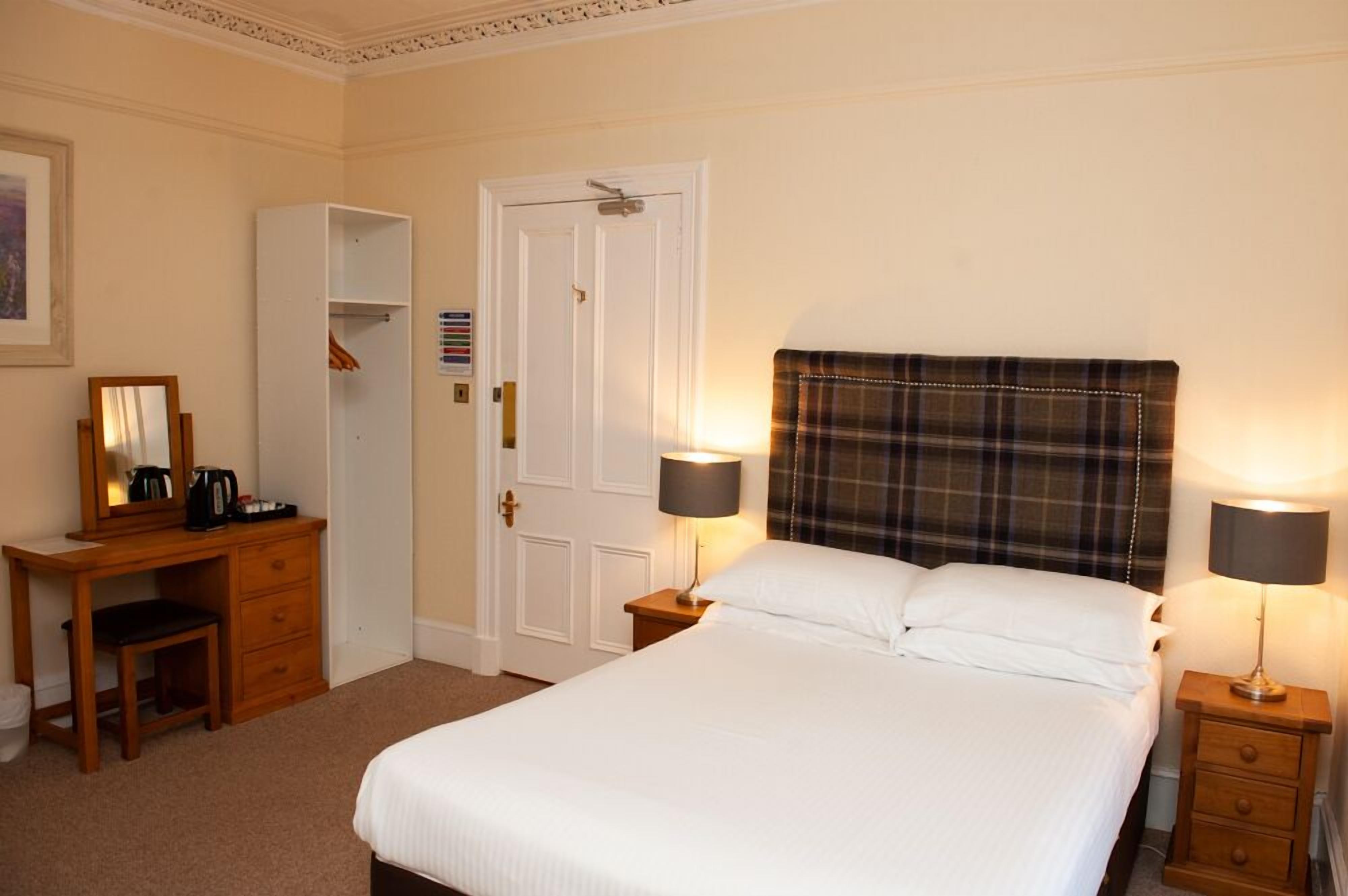 A neatly arranged hotel room with a bed, bedside lamps, a desk with a chair, a wardrobe, and a door.