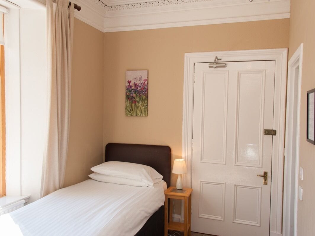 A hotel room with a bed, a nightstand with a lamp, a door, and a wall-mounted painting.