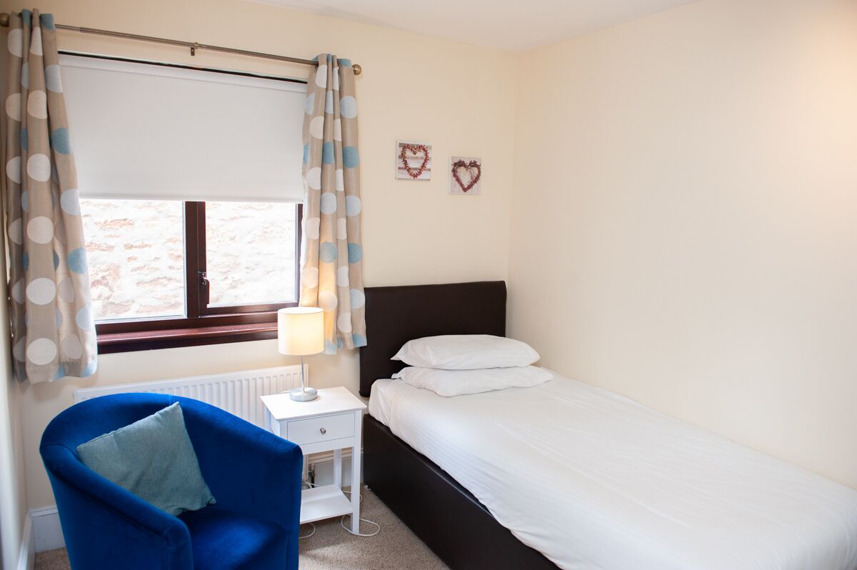 Un dormitorio con una cama individual, una mesita de noche, una lámpara, una silla azul y una ventana con cortinas.