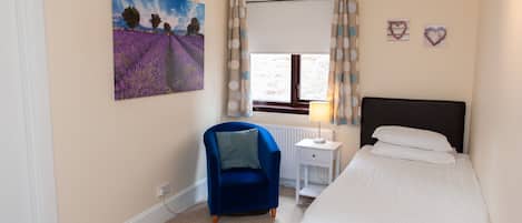 Ein Schlafzimmer mit Bett, Sessel, Heizkörper, einem Fenster mit Vorhängen und einem Bild an der Wand.