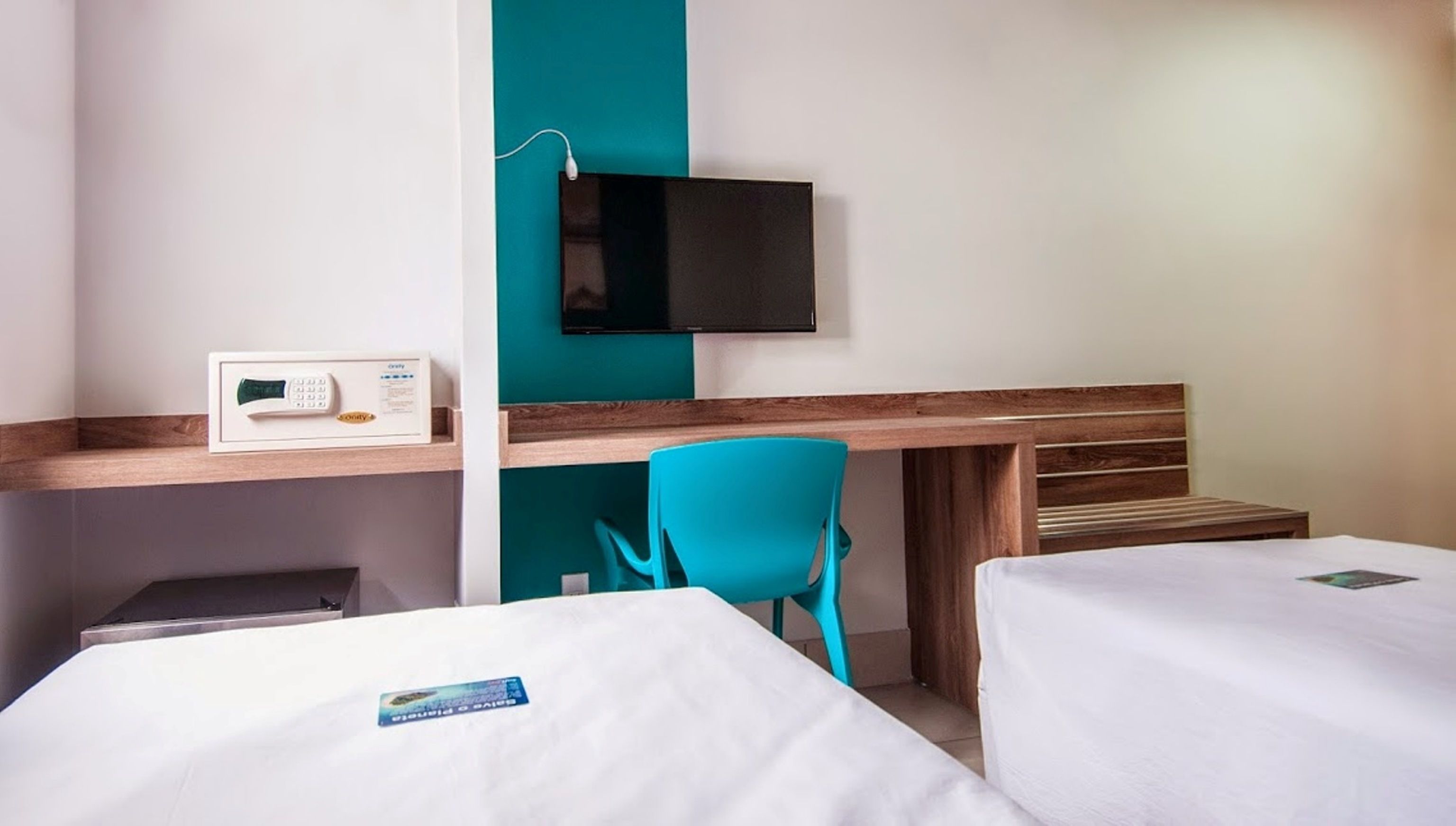 A hotel room with two beds, a desk with a computer, a teal chair, and a flat-screen TV mounted on the wall.