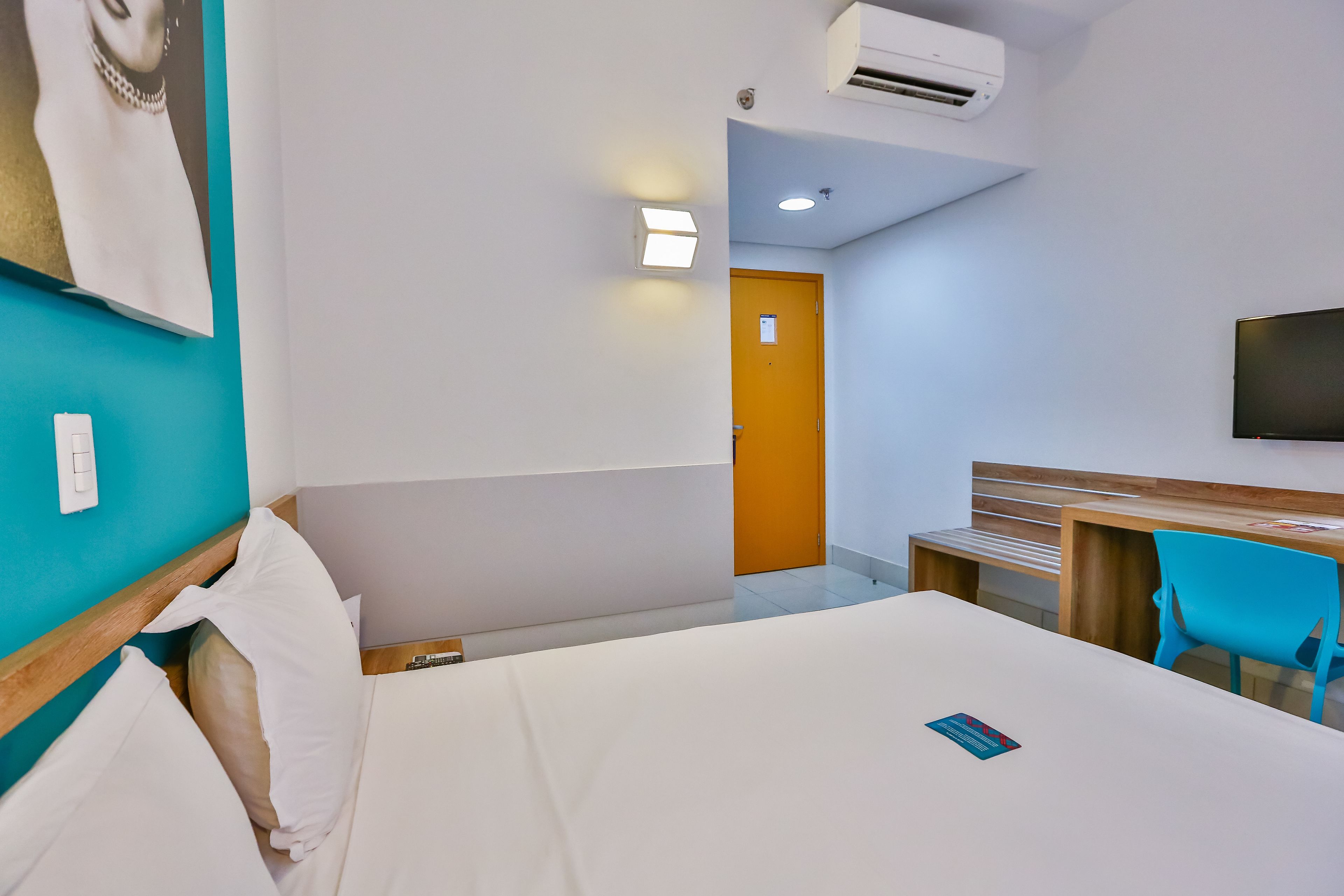 A hotel room with a bed, a desk, a chair, a TV, and an air conditioning unit.