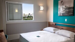 A bedroom with a bed, a window with blinds, a city view, a teal accent wall, and a framed picture.