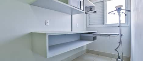 Un baño moderno y limpio con paredes blancas, un mueble estante empotrado, una ventana con persianas y una aspiradora de pie.