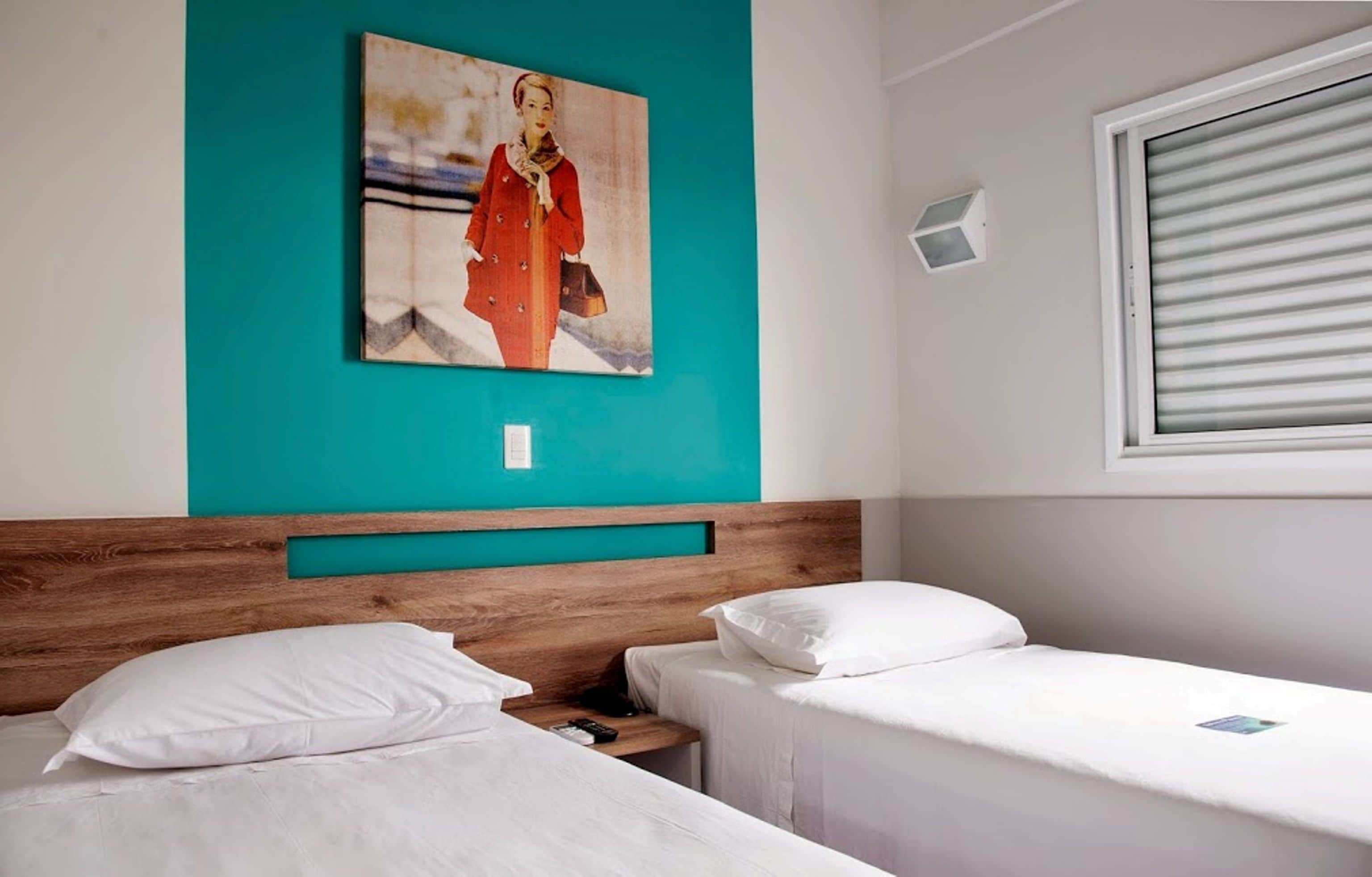 Una habitación de hotel con dos camas individuales, un cabecero de madera, una pared de acento color aguamarina, un cuadro, una ventana con persianas y una mesita de noche pequeña.