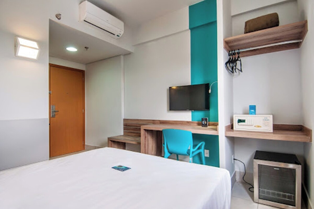 A hotel room with a bed, a desk, a chair, a TV, a heater, and a shelf with a microwave.