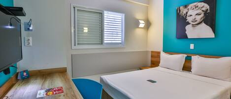 A hotel room with a bed, a desk, a chair, a TV, a window with blinds, and a wall-mounted picture.