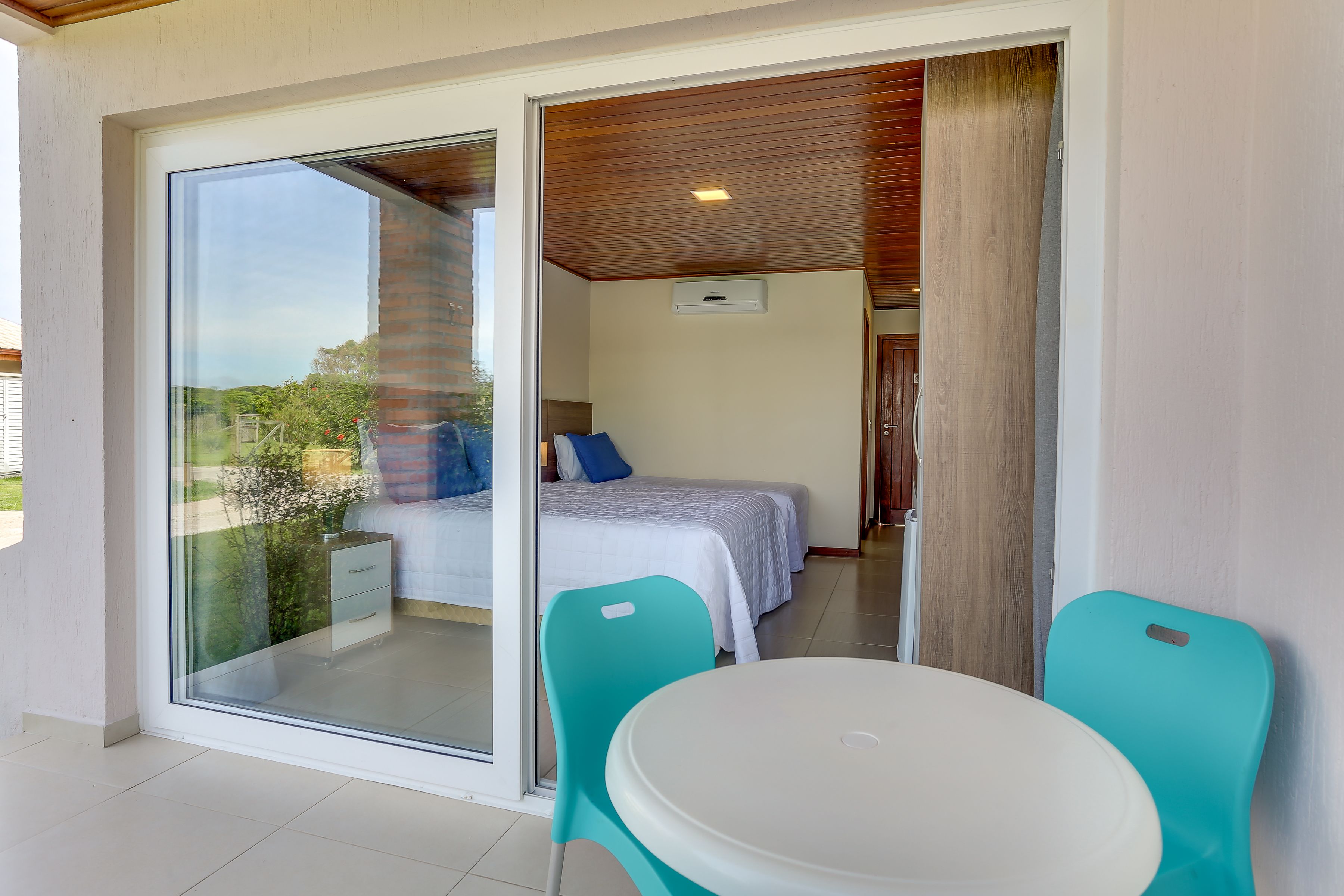 Una habitación con una puerta de cristal corredera que da a un balcón, una cama con ropa de cama blanca y almohadas azules, y una pequeña mesa con dos sillas turquesas.