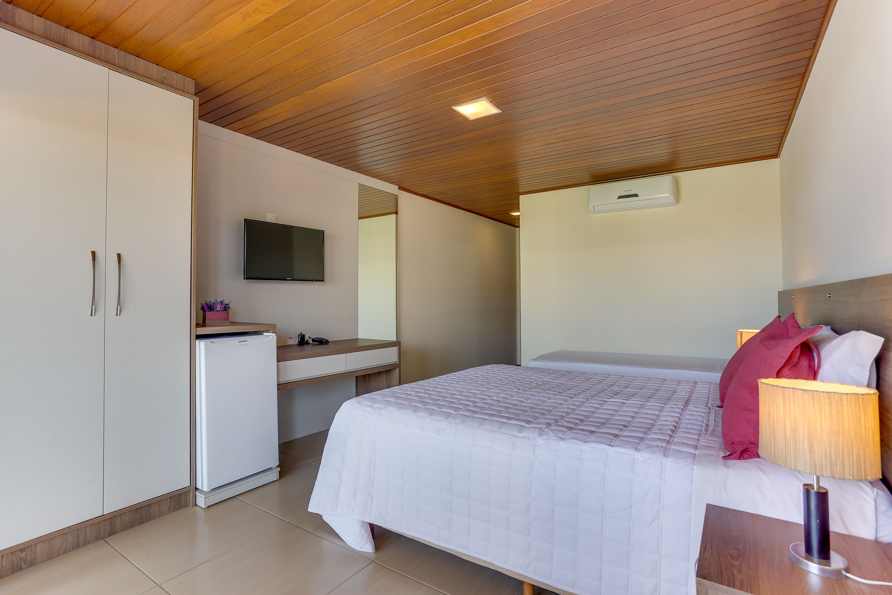 Quarto com cama, mesa de cabeceira, escrivaninha, guarda-roupa e televisão fixada na parede.