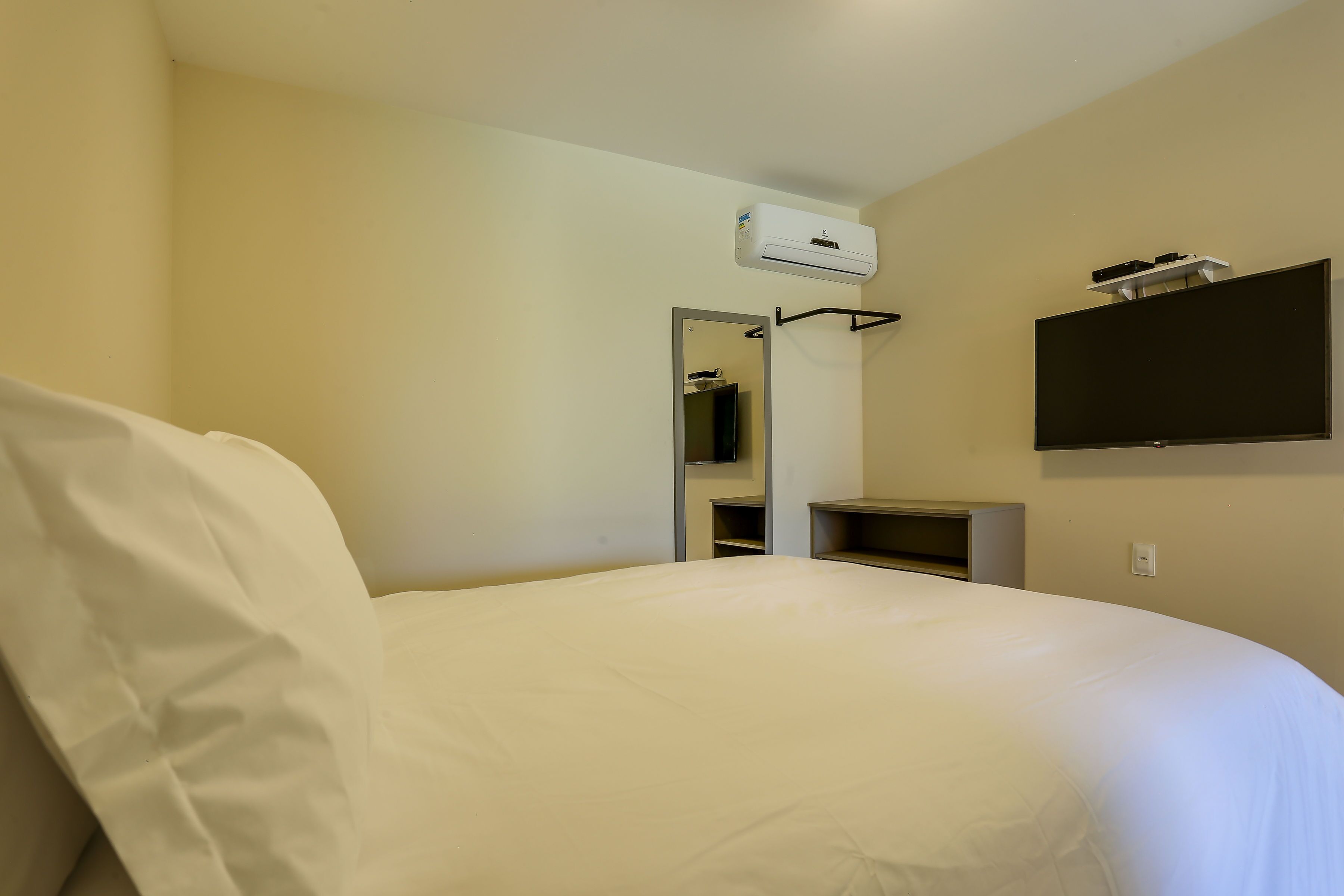 A hotel room with a bed, a TV mounted on the wall, a mirror, and a small shelf.