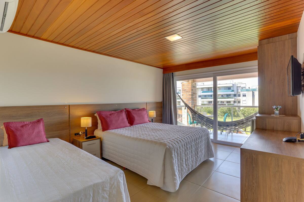 A hotel room with two beds, a wooden ceiling, a balcony with a hammock, and a TV on a wooden stand.