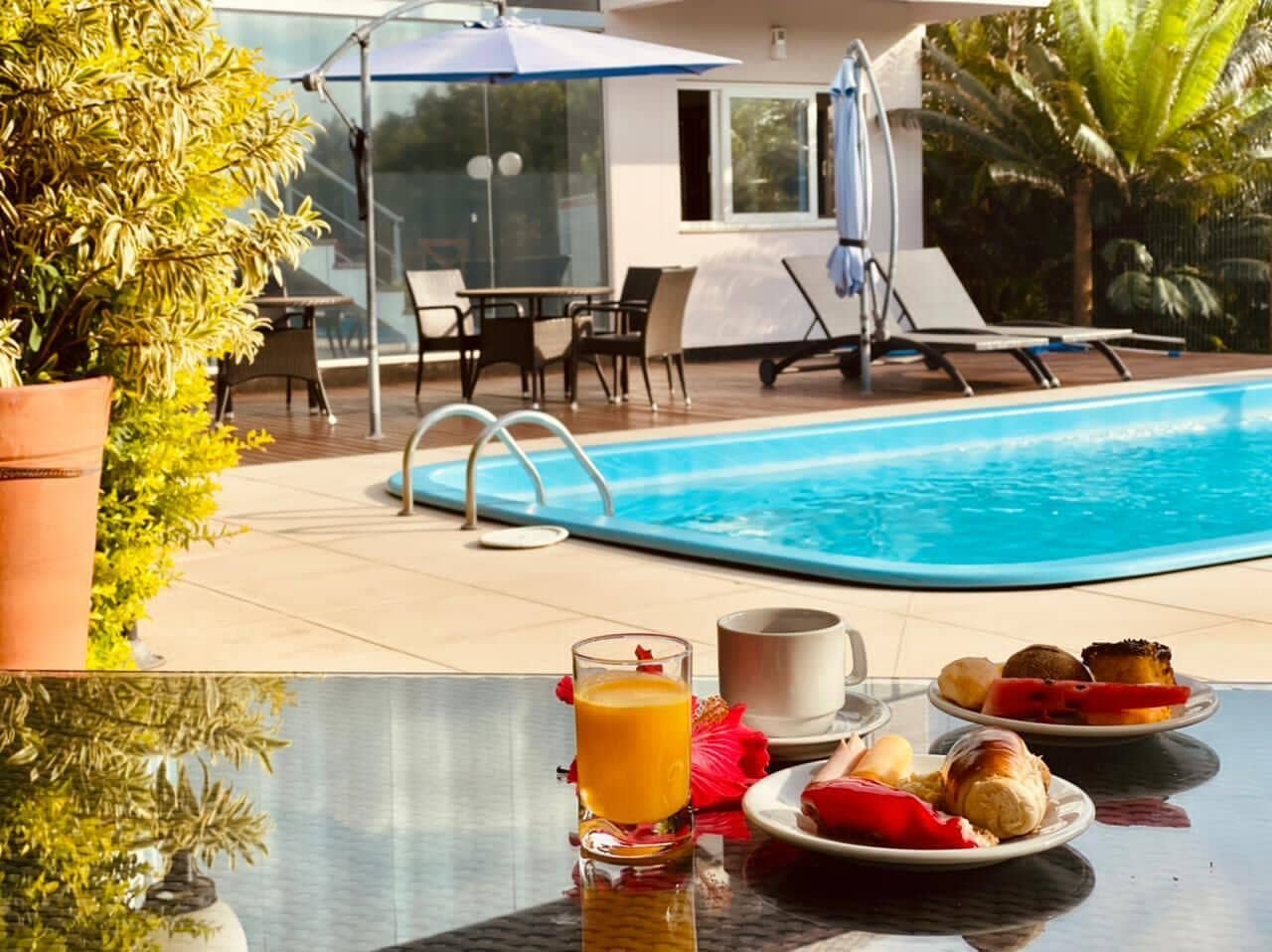 Un petit-déjeuner servi au bord de la piscine, avec un verre de jus d’orange, une tasse et des assiettes remplies de nourriture.
