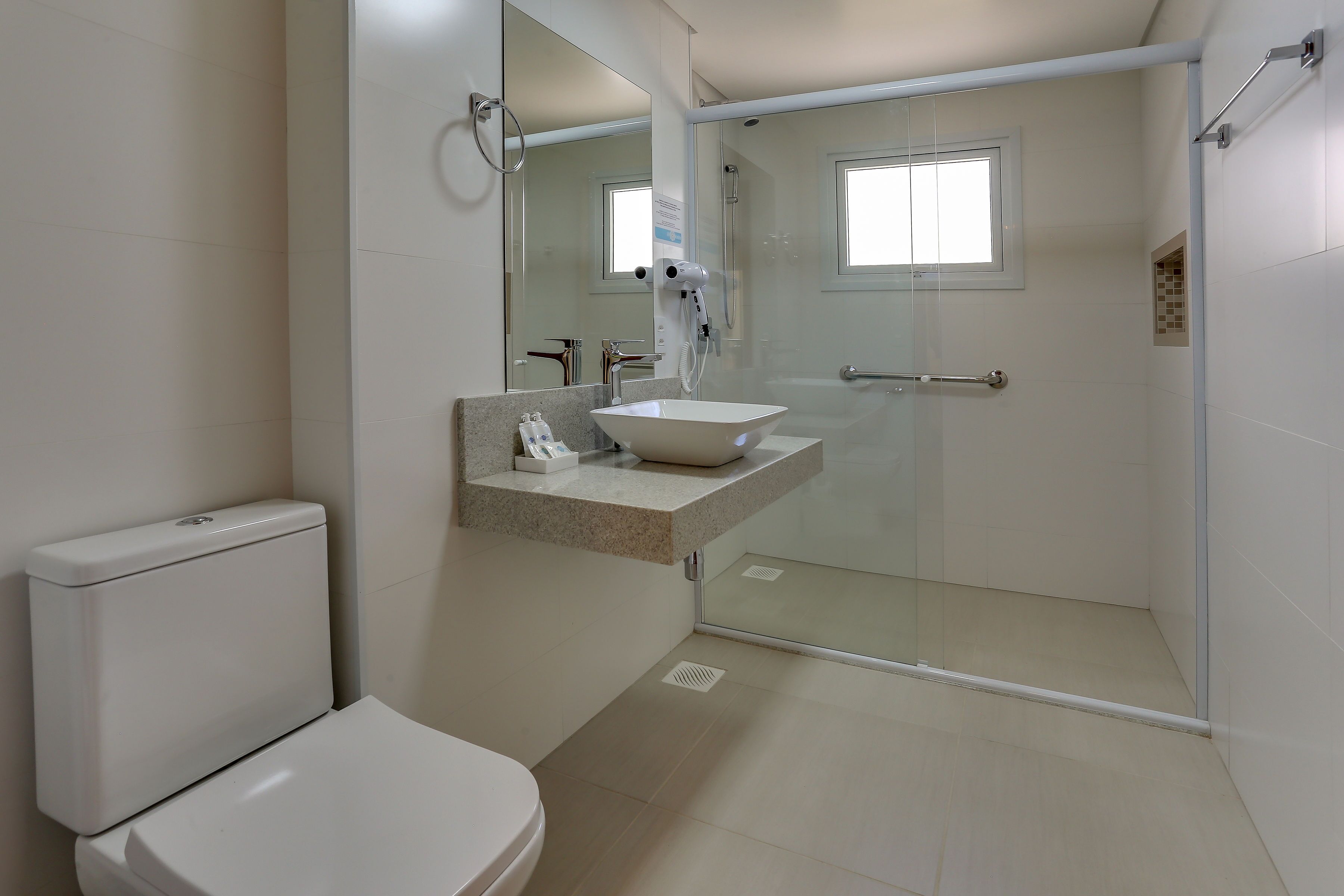 Une salle de bain moderne comprenant des toilettes, un lavabo avec un meuble-vasque, une douche en verre et une fenêtre laissant entrer la lumière naturelle.