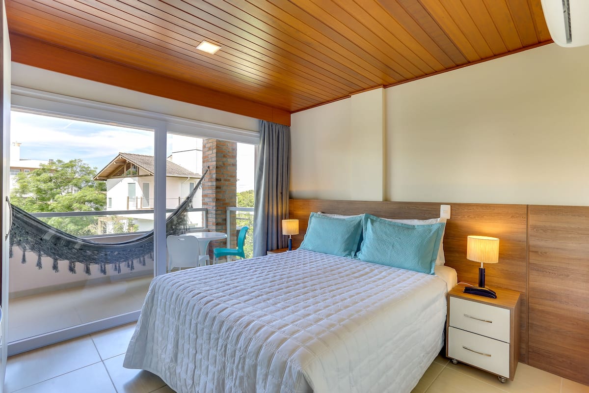 A bedroom with a large bed, a nightstand with a lamp, a wooden ceiling, and a view of a balcony with a hammock.