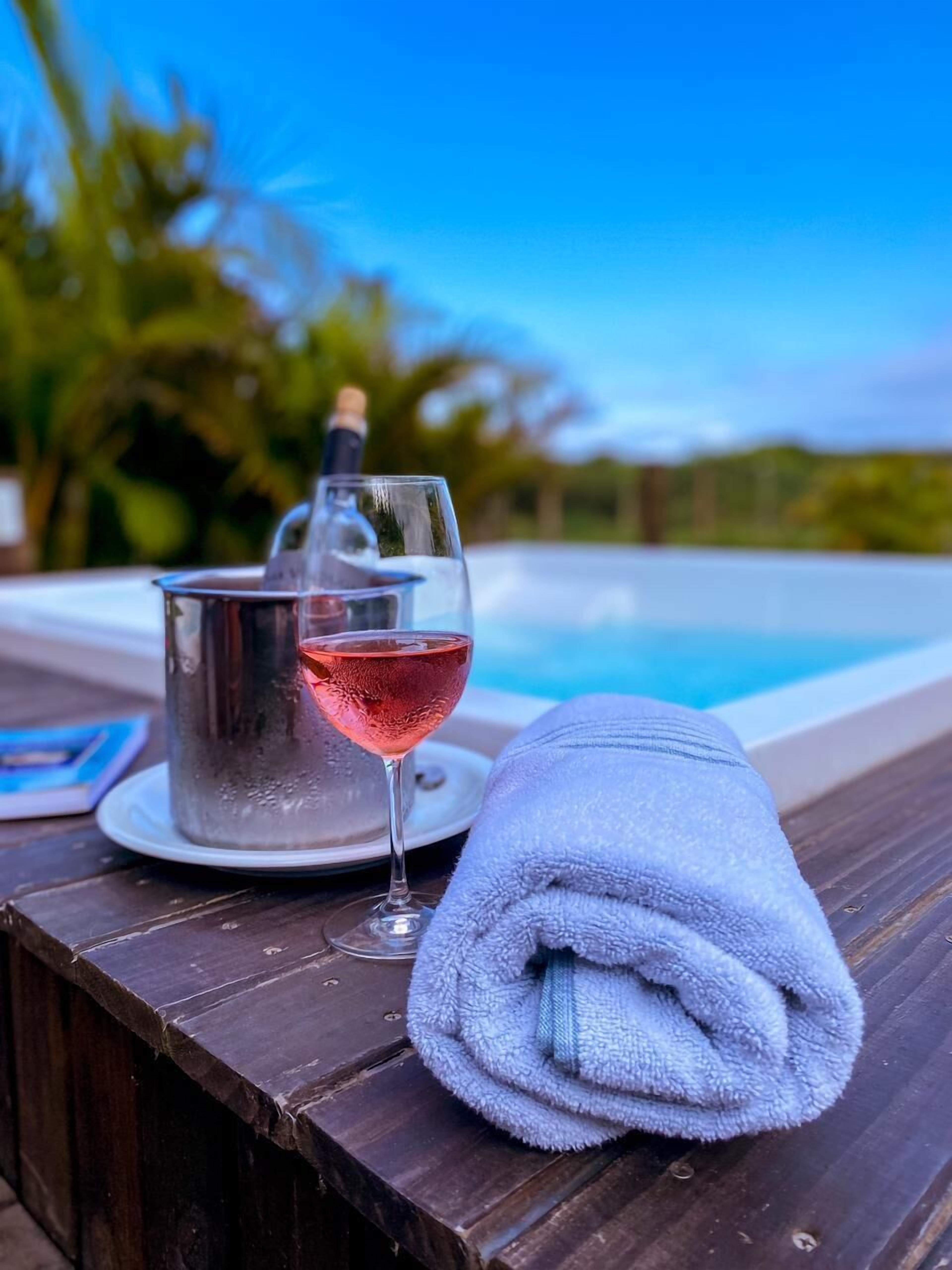 Taula de fusta amb un got de vi negre, una ampolla de xampany i una tovallola doblegada al costat d'una piscina.