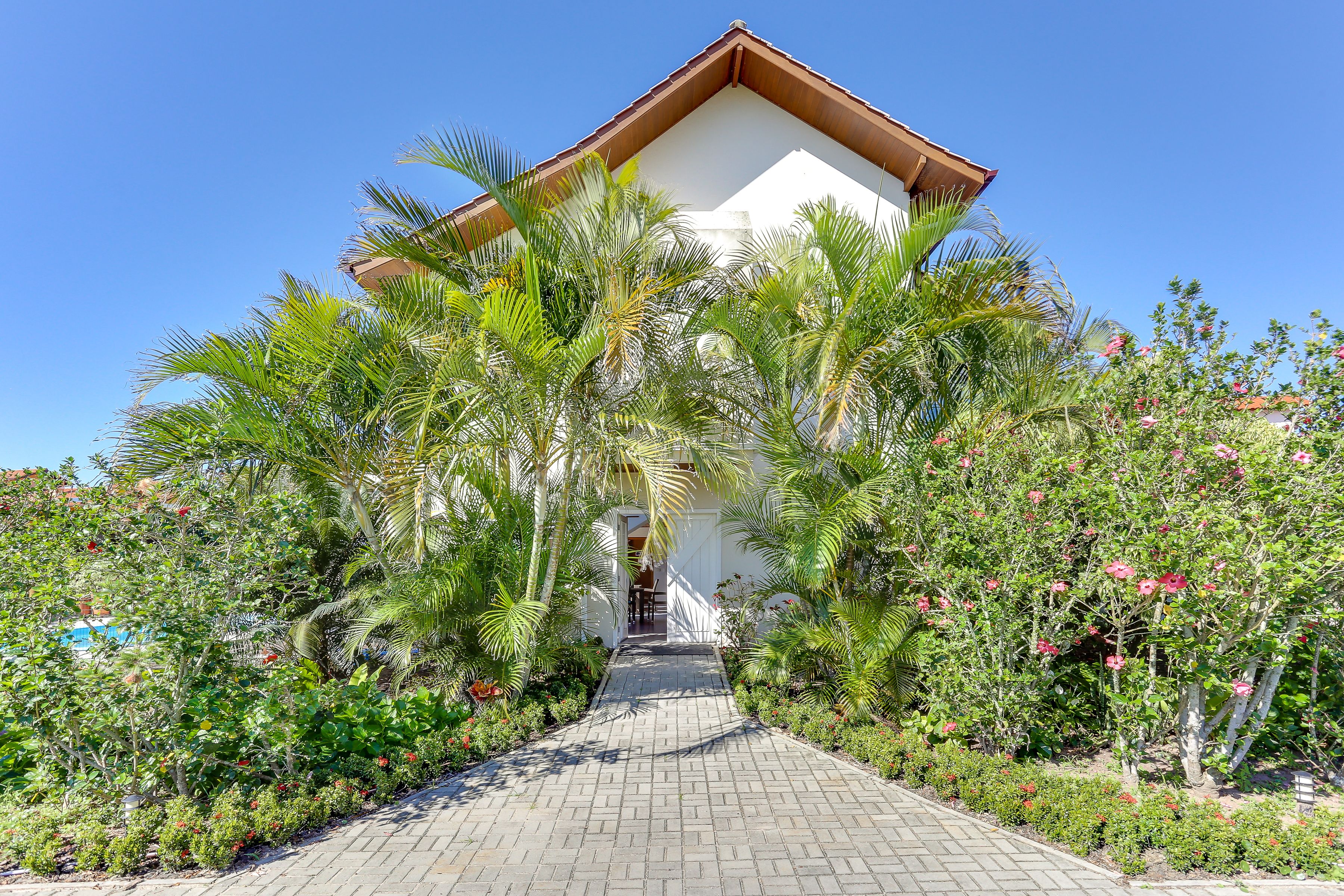 A pathway leading to a building with a white facade and a brown roof, surrounded by lush greenery and flowering plants.