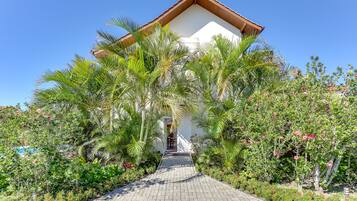 A pathway leading to a building with a white facade and a brown roof, surrounded by lush greenery and flowering plants.