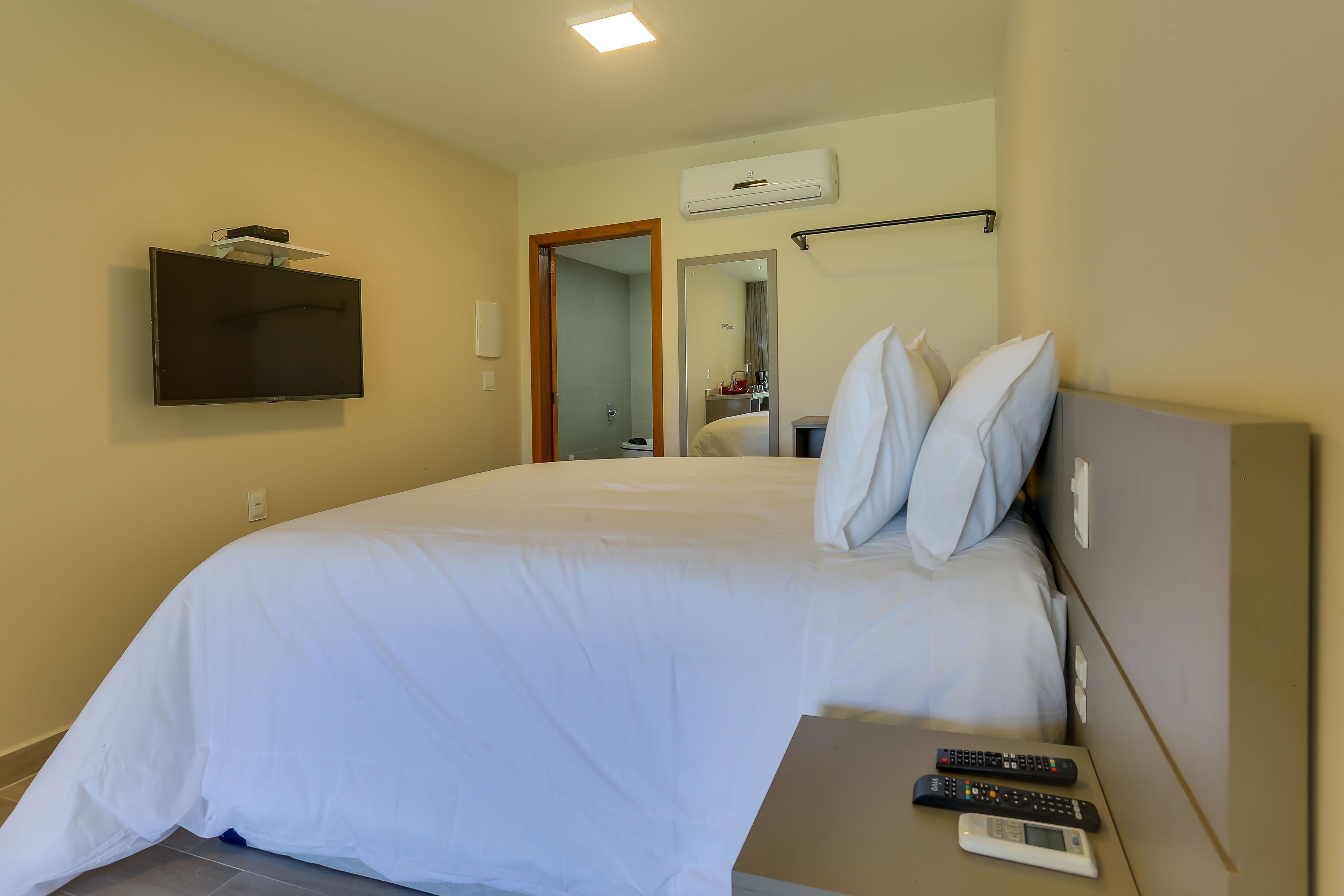 A hotel room with a large bed, a TV mounted on the wall, a bedside table with a remote control, and an air conditioning unit above the door.