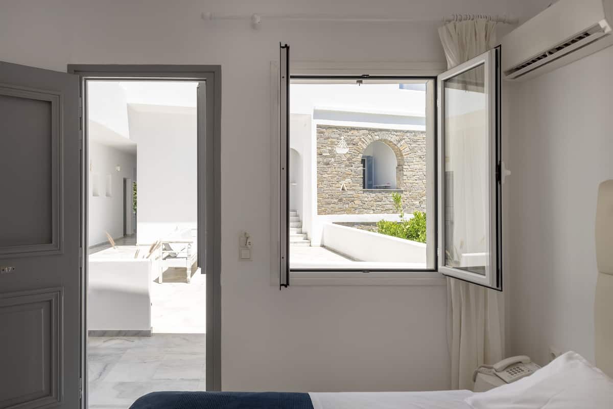 A hotel room with a bed, a telephone, and a window with a view of a courtyard and a stone building.