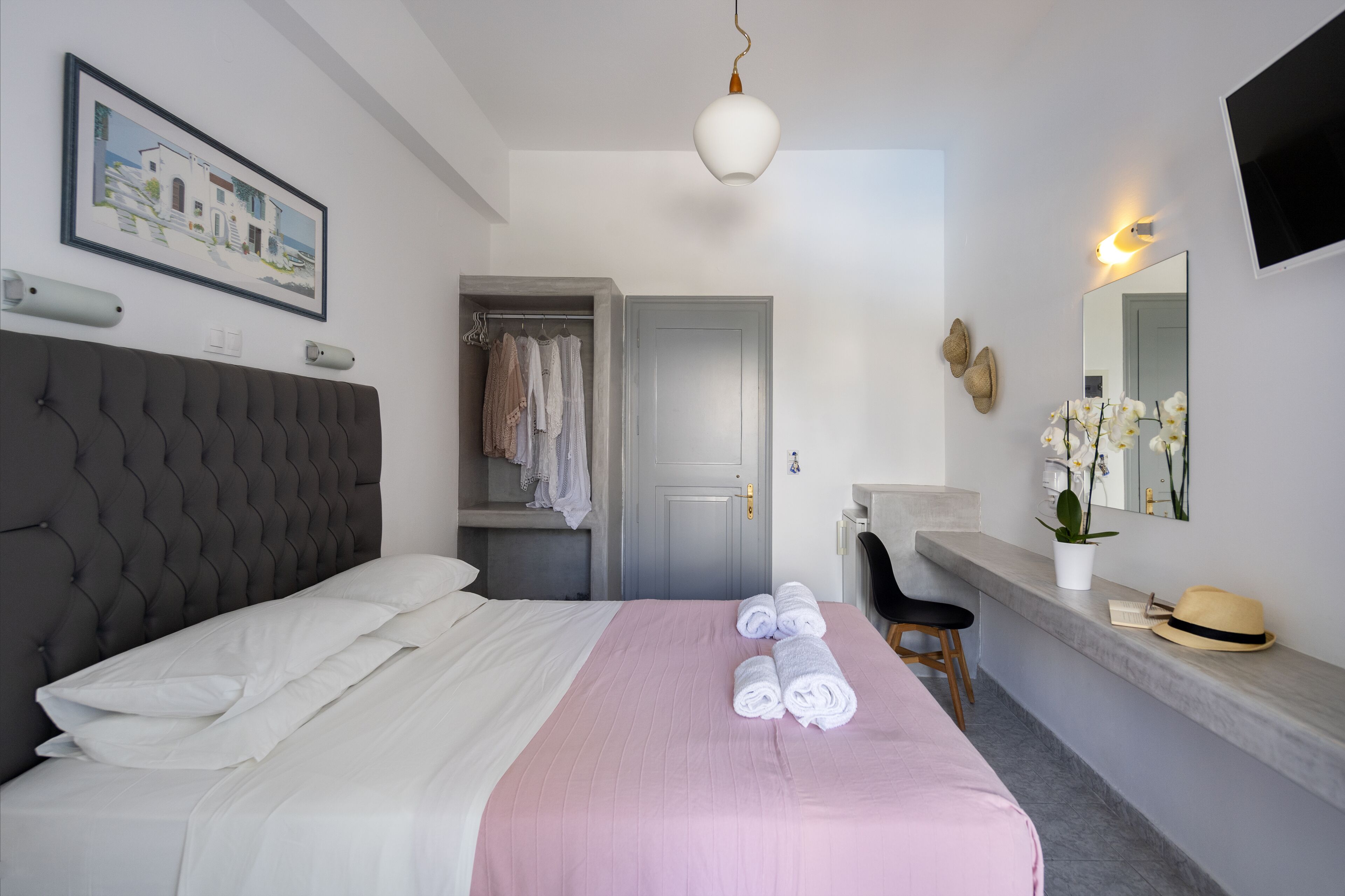 A neatly made bed with white linens, a pink bedspread, and rolled towels. There is a grey upholstered headboard, a grey door, and a small seating area with a chair.