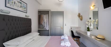 A neatly made bed with white linens, a pink bedspread, and rolled towels. There is a grey upholstered headboard, a grey door, and a small seating area with a chair.