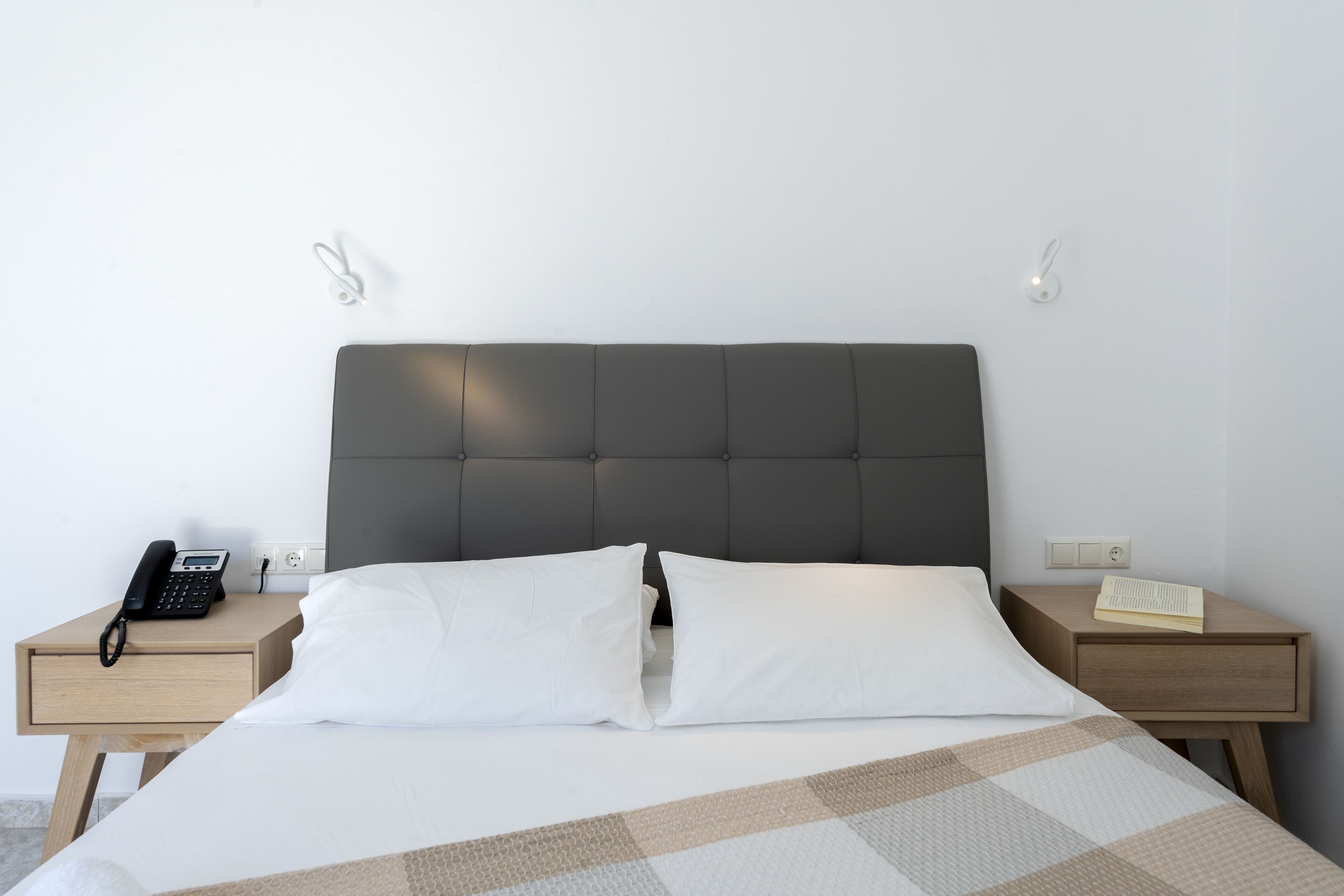 A neatly made bed with white pillows and a checkered blanket, a bedside telephone, and a small wooden nightstand with a book.