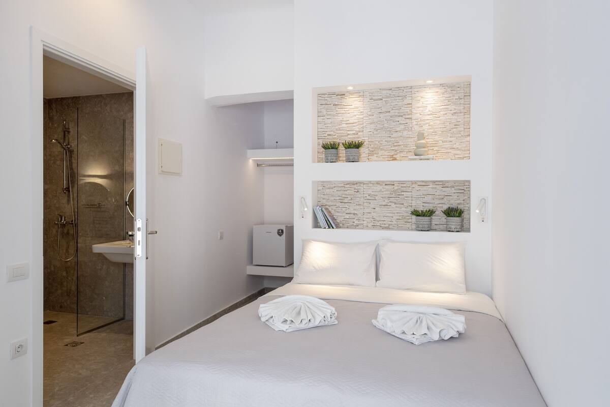 A modern hotel room with a shower, a bed with neatly folded towels, and decorative wall shelves with plants.