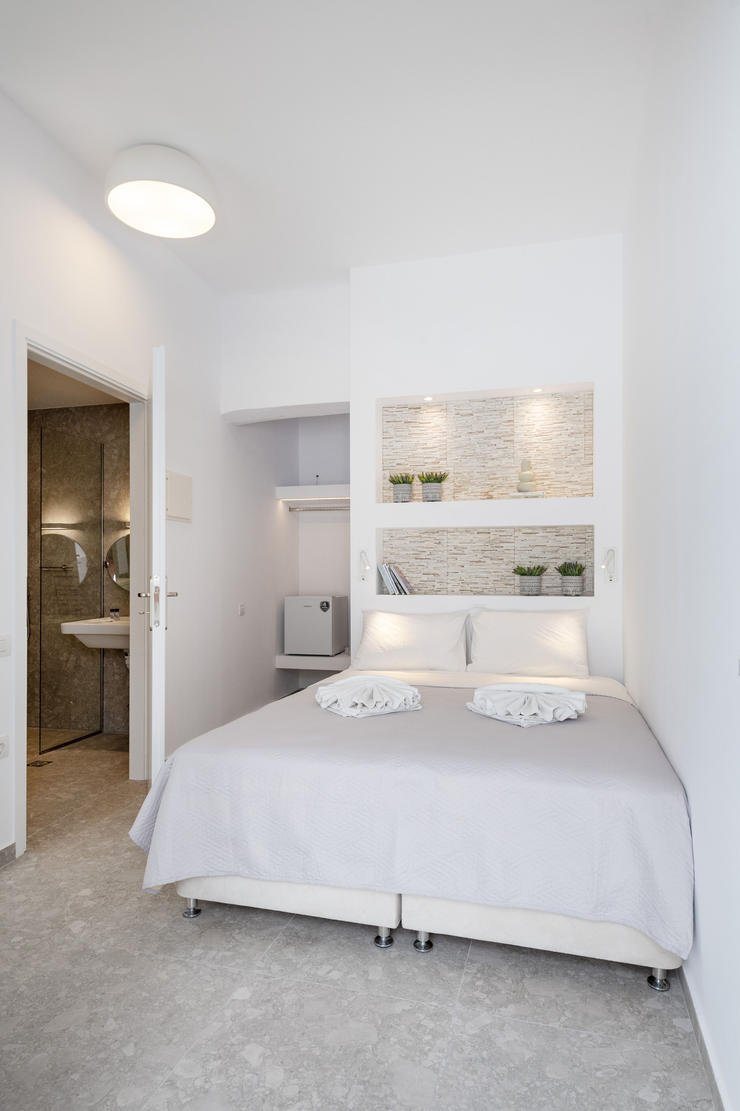 Ein modernes Schlafzimmer mit einem Bett, Nachttischen, einer Wandablage mit Pflanzen und einem durch eine offene Tür einsehbaren Badezimmer.