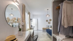 Una habitación de hotel moderna con un espejo redondo grande, una orquídea en maceta, un sombrero amarillo, una cama con colcha azul y un perchero con ropa colgada.