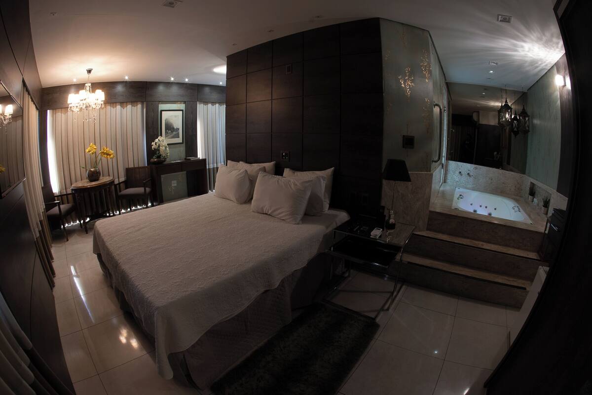 A modern hotel room with a large bed, a sitting area with a table and chairs, a small desk, a vanity with a mirror, and a separate bathroom with a bathtub.