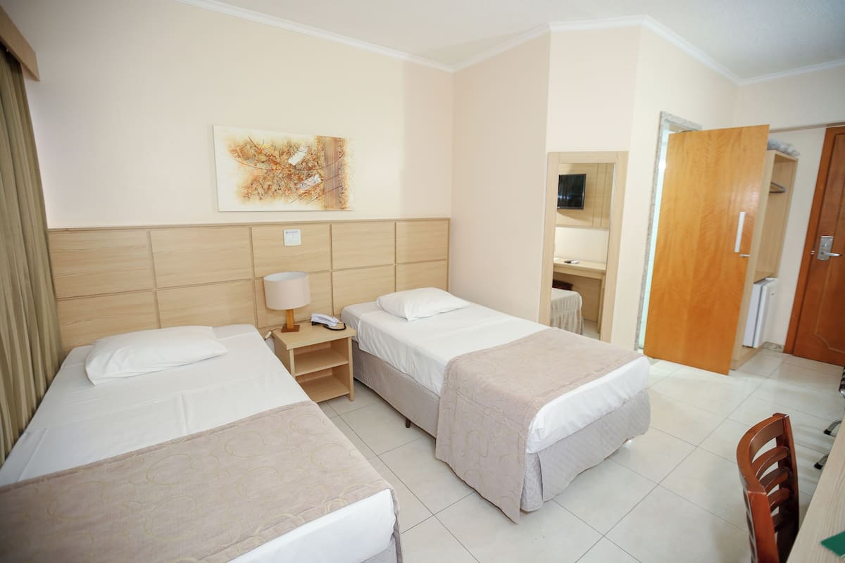 A hotel room with two single beds, a bedside table with a lamp, a wall-mounted artwork, and a wooden door.