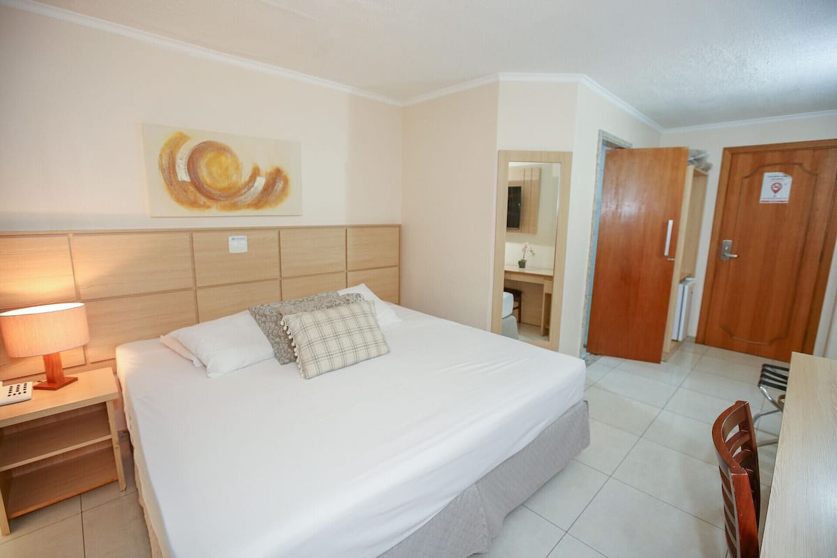 A hotel room with a bed, a bedside table with a lamp, a mirror, a wooden door, and a bathroom visible through an open door.