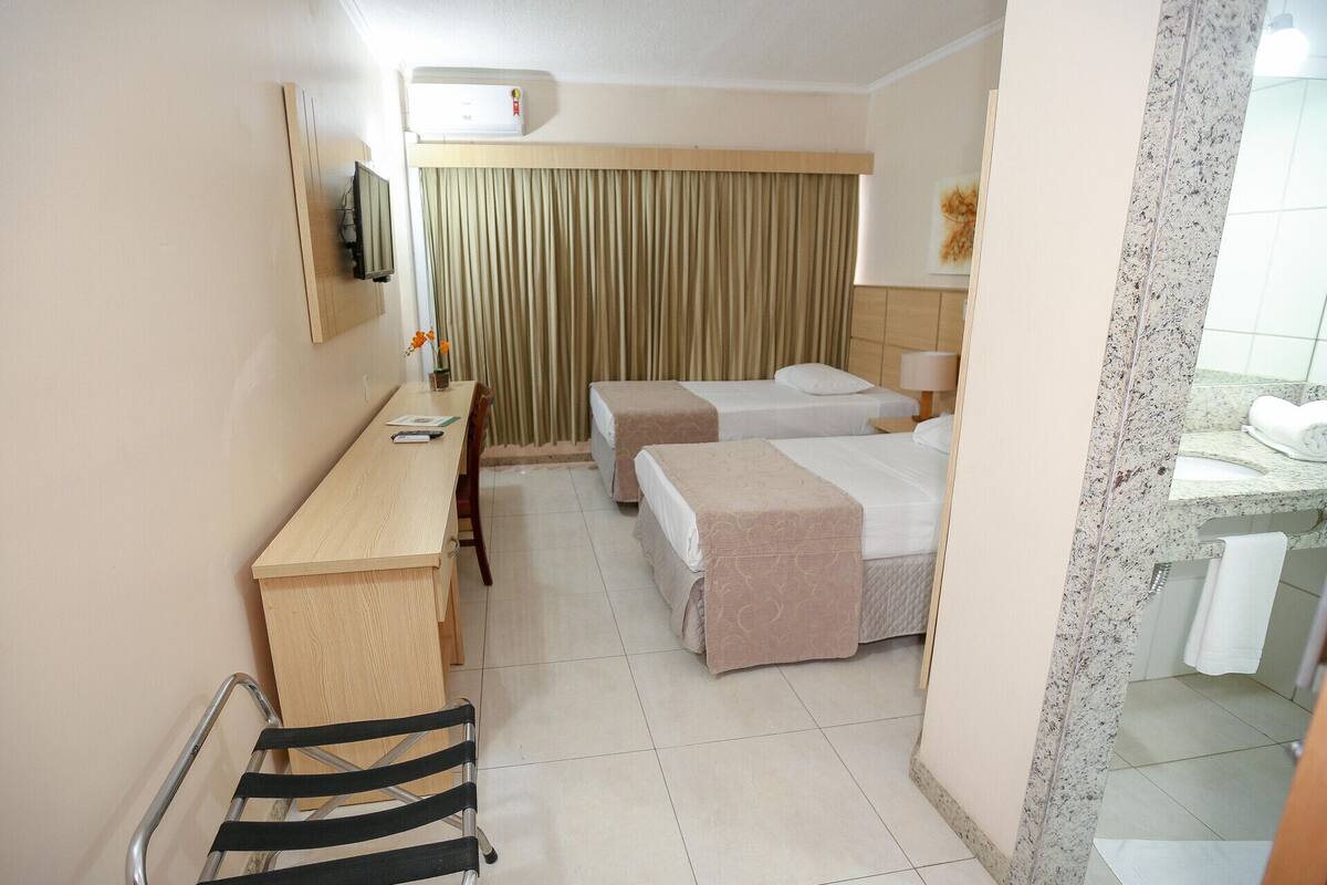 A hotel room with a desk, two beds, a TV, an air conditioner, and a bathroom.