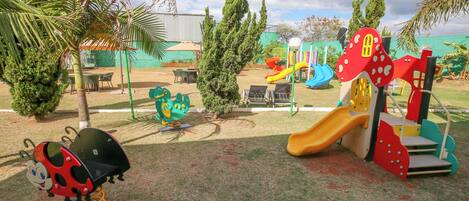 A playground with slides and climbing structures, surrounded by trees and a building in the background.
