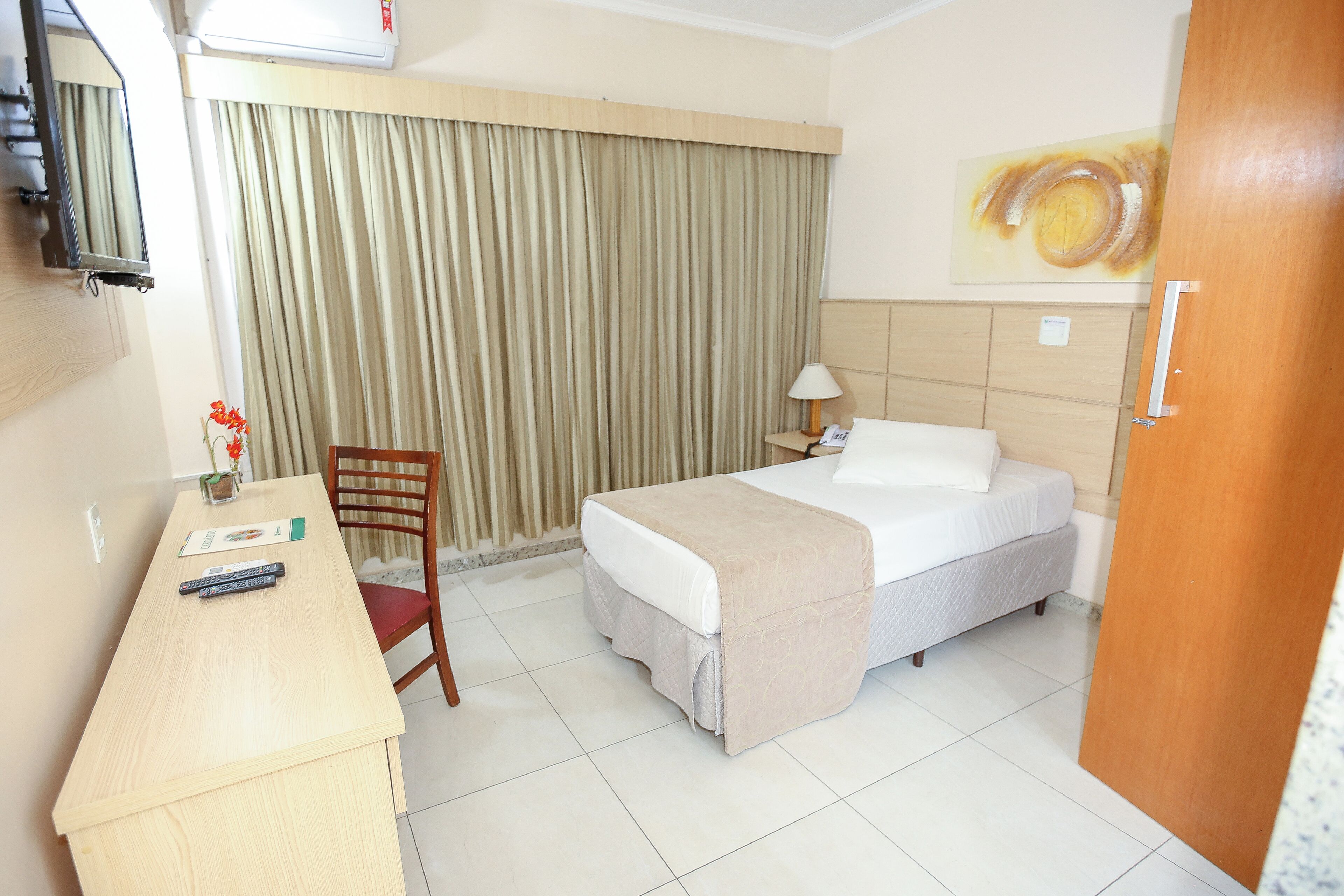 Quarto de hotel com cama, escrivaninha, cadeira, abajur e um quadro na parede.