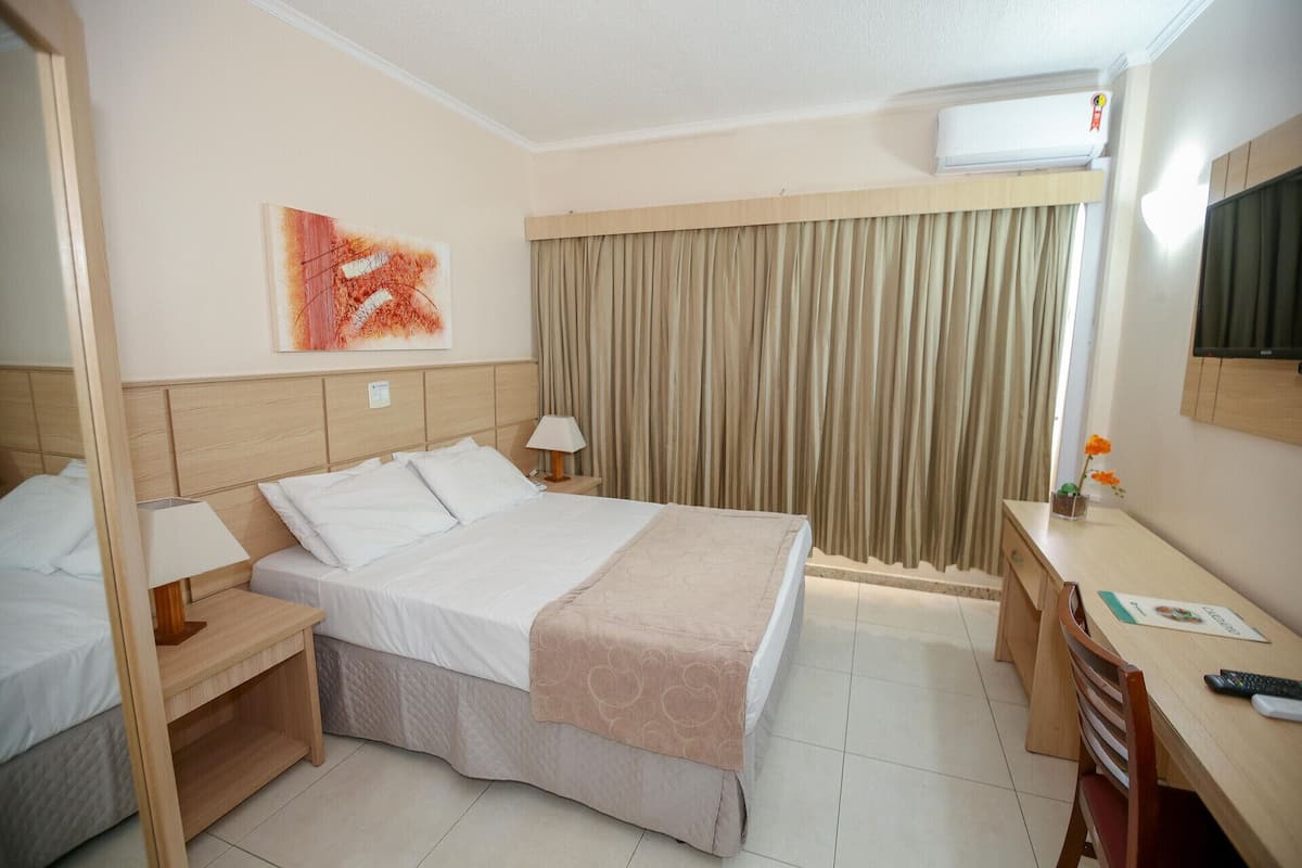 A hotel room with a bed, bedside table, lamp, desk, chair, and an air conditioning unit.