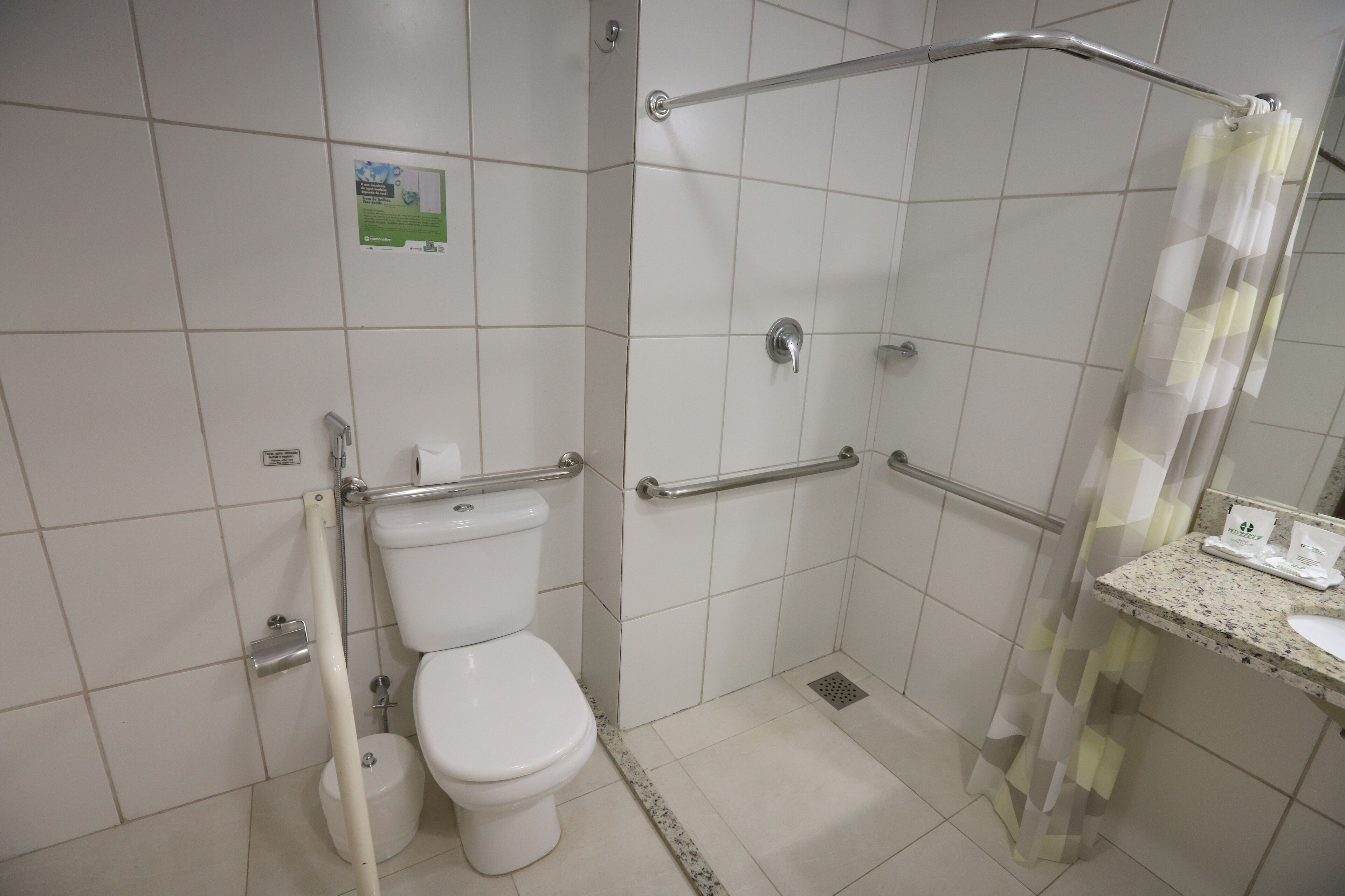 Un baño con inodoro, barras de apoyo, ducha con puerta de vidrio y lavamanos con espejo.