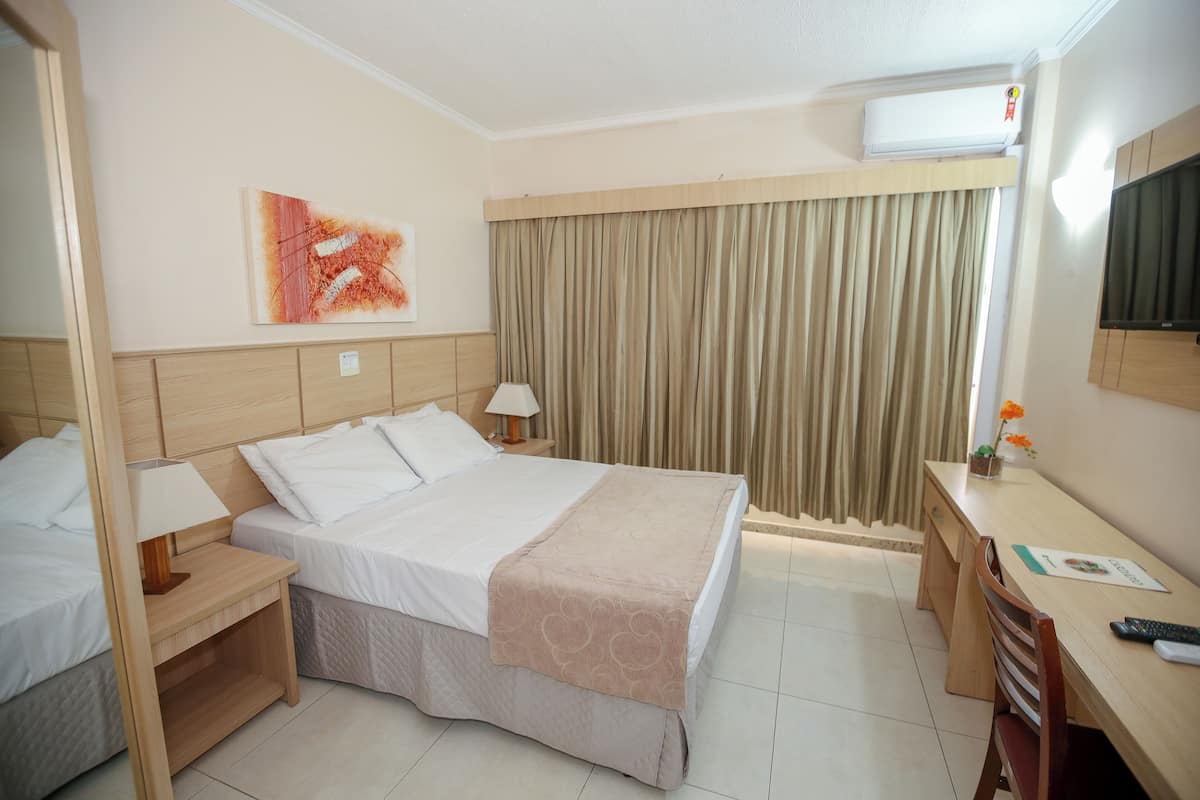 A hotel room with a bed, bedside tables, a desk, a chair, a TV, and a window with curtains.