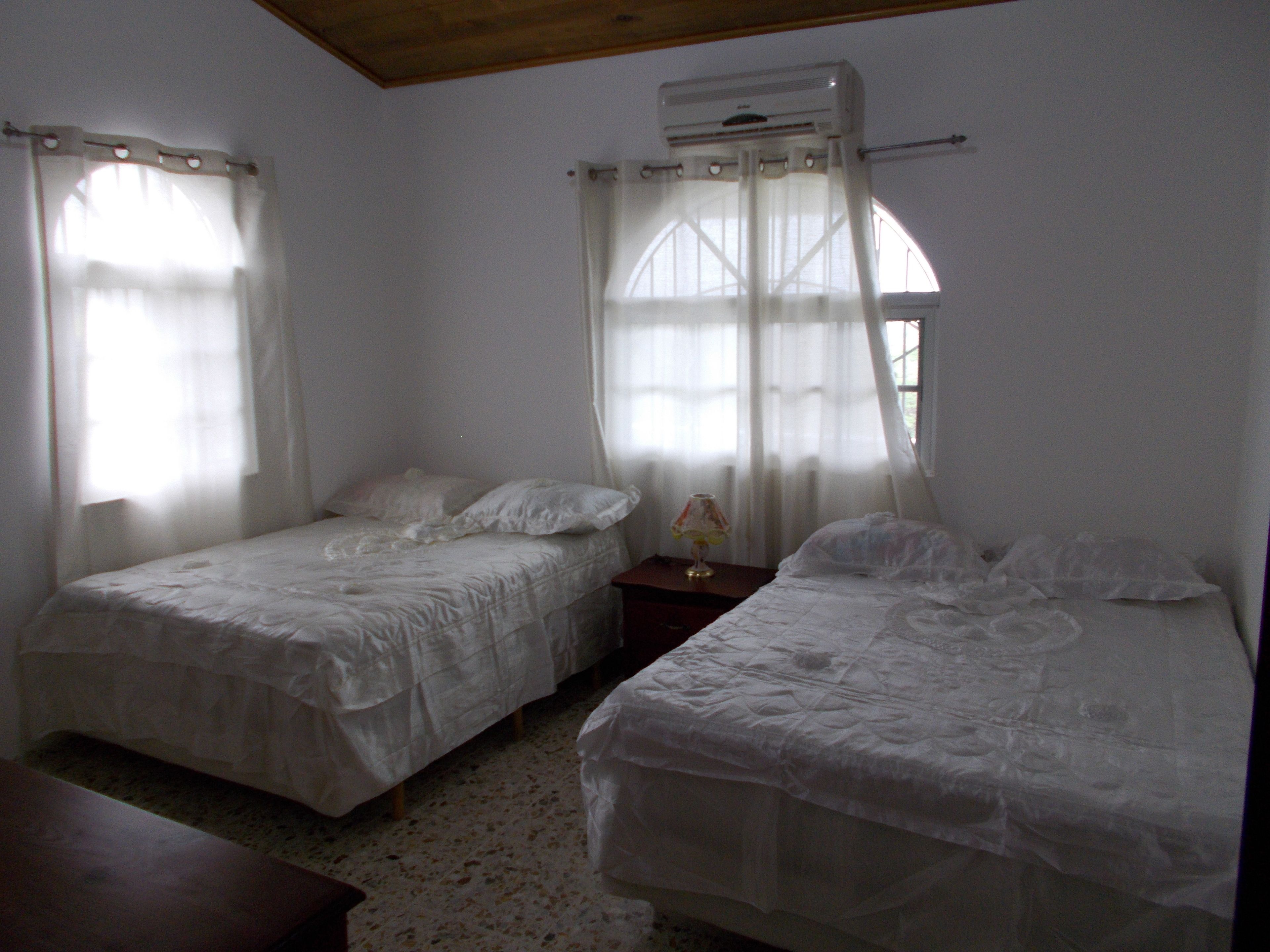A room with two single beds, a wooden headboard, a small table with a lamp, and a window with curtains.