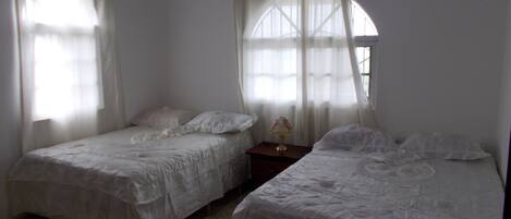 A room with two single beds, a wooden headboard, a small table with a lamp, and a window with curtains.