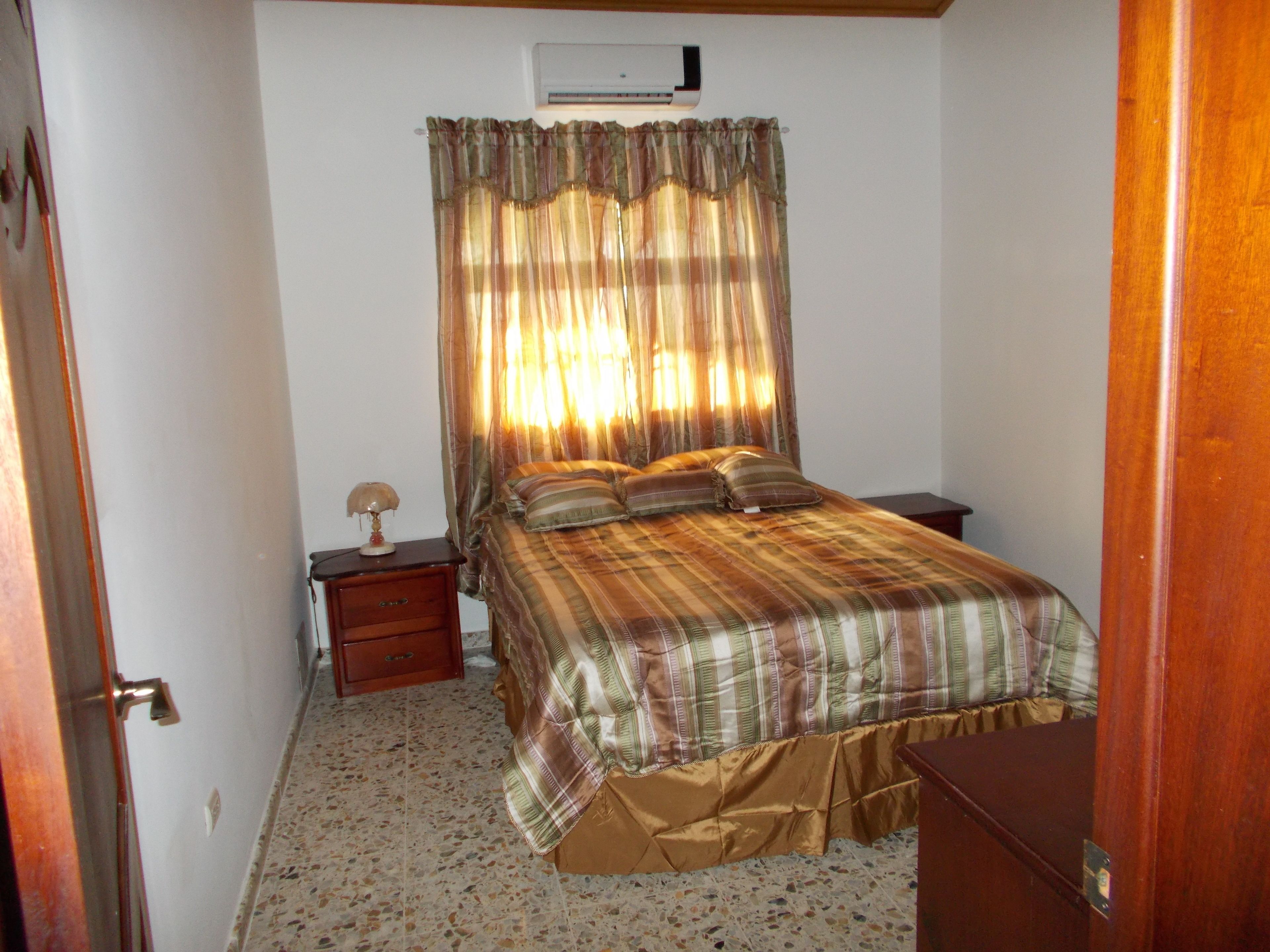 A bedroom with a bed, a nightstand, a lamp, and an air conditioning unit on the wall.