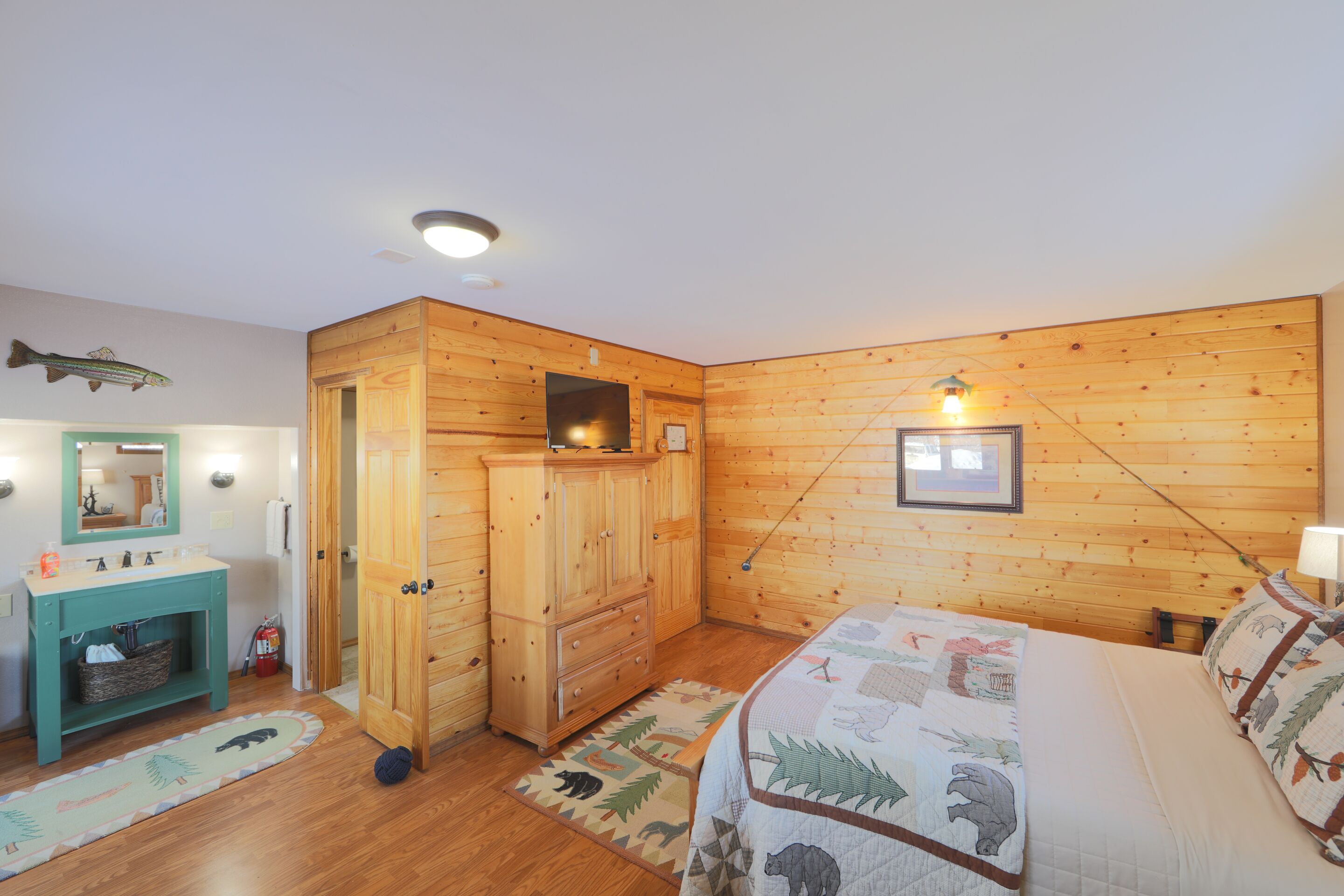 A room with wooden walls, a bed with a quilt, a wooden wardrobe, a sink with a mirror, and a rug.